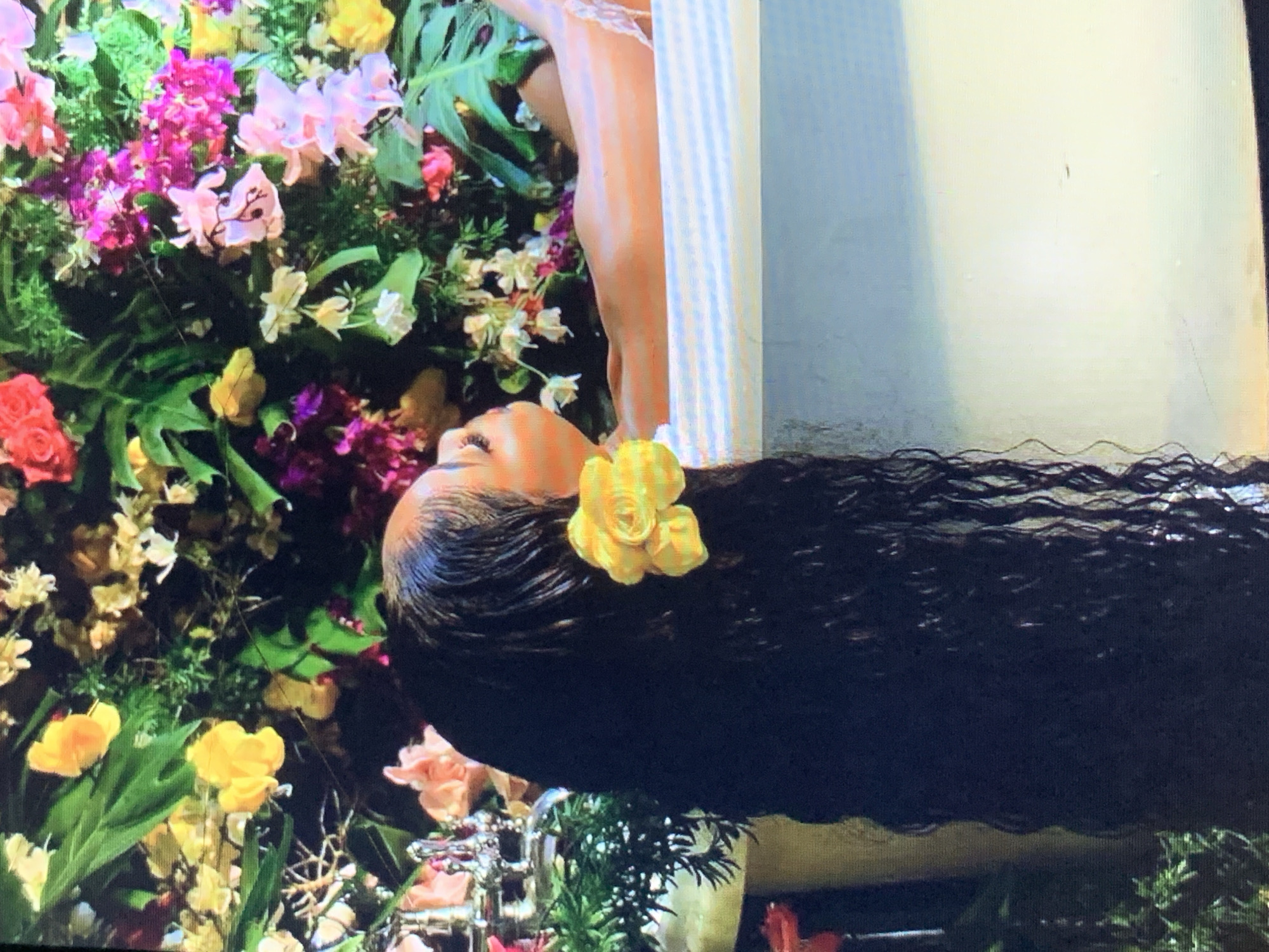 A woman with dark, long, wavy hair is lying among a variety of colorful flowers. A yellow flower is placed in her hair. A portion of the woman's body, extending from her shoulder to her thigh, is visible. Her body is positioned alongside a white, vertical object. The environment around her is filled with a dense arrangement of flowers in shades of pink, yellow, and white, interspersed with green foliage. A silver object is situated near the bottom of the frame, partially obscured by greenery. The scene is indoors. The location of the scene is not confirmed but is said to be inside the New Stadium in Nigeria.