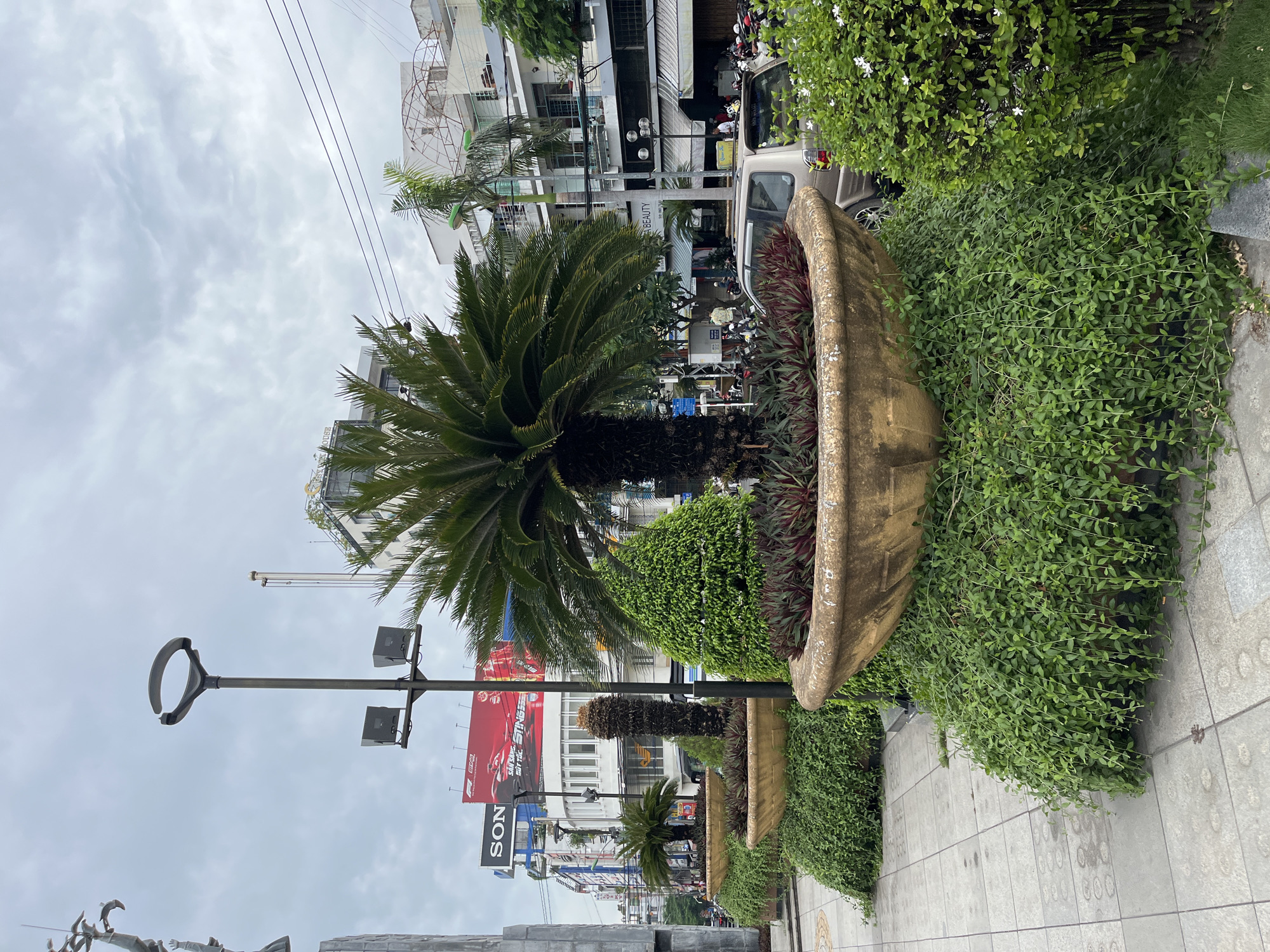 The image shows a street scene in Nha Trang, Vietnam, with a prominent series of large, decorative planters filled with lush greenery and palm-like plants. A tall street lamp with two speakers is positioned to the left of the central planter. In the background, buildings line the street, some featuring awnings and signage, including a large red billboard with "SONY" and "STING" text. A silver SUV is parked on the street behind the planters, with numerous motorbikes also visible, suggesting a busy urban environment. The sky is overcast, indicating a cloudy day. The foreground shows a tiled sidewalk and the base of the planters with abundant trailing green foliage.