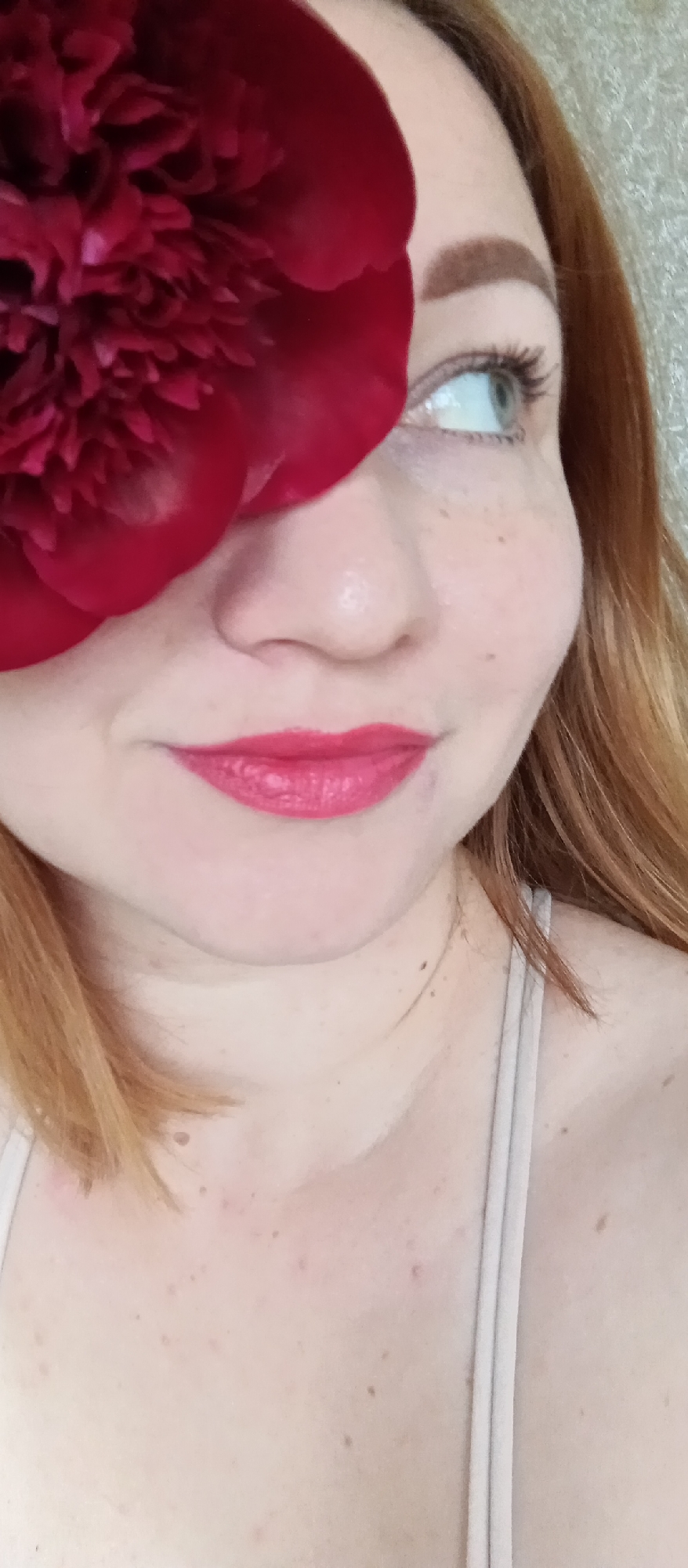 The image depicts a close-up of a fair-skinned woman with red hair. Her face is partially obscured by a large, dark red flower, possibly a peony. She has freckles and is wearing pink lipstick. Her eye is visible, looking upward. A thin-strapped top is visible at her shoulder and chest. The background is a textured off-white. The location is not specified in the image.