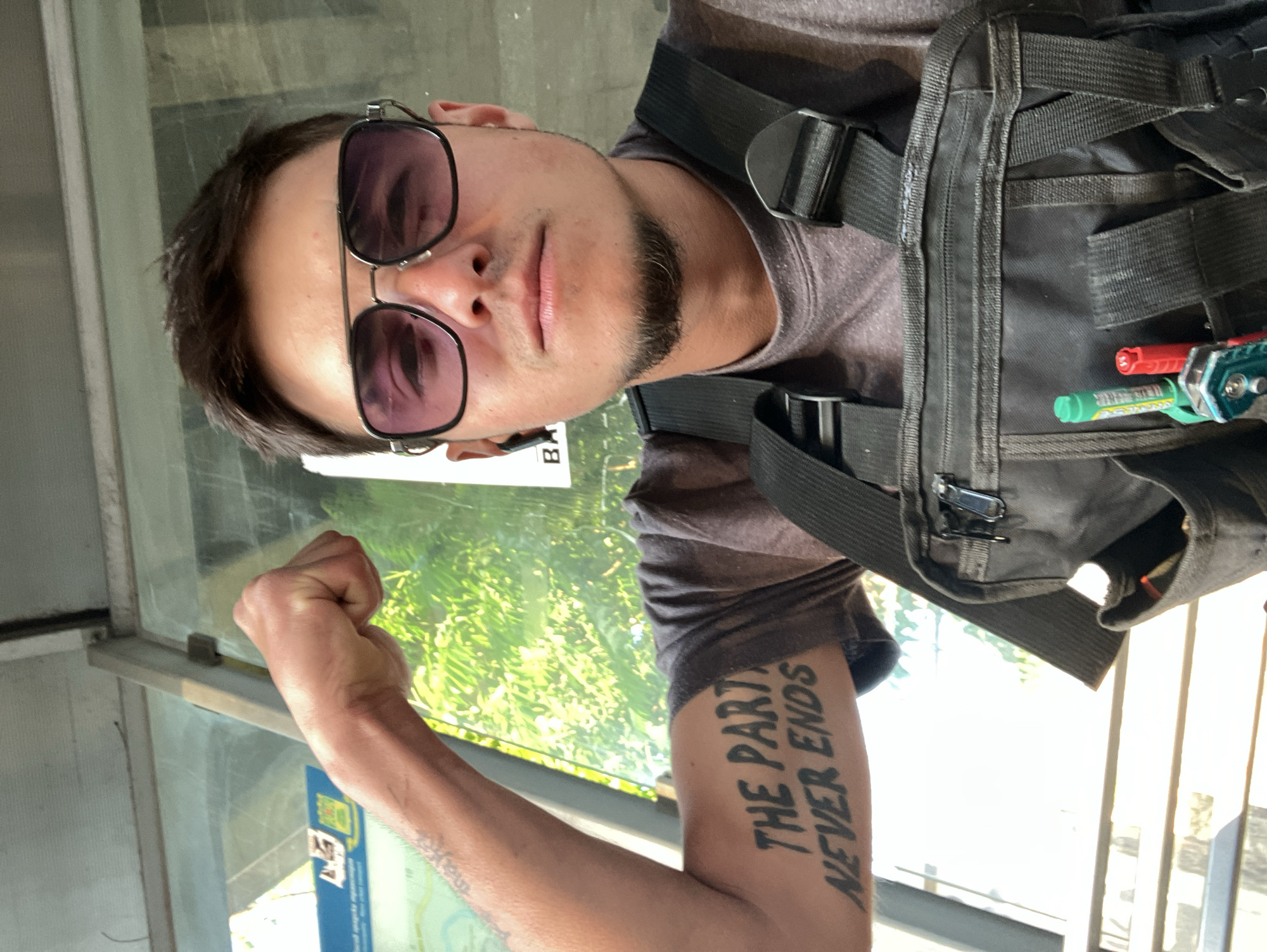 A selfie depicts a man with short dark hair, a beard, and sunglasses. He is wearing a dark gray t-shirt and a black tactical vest with straps and buckles. He holds a clenched fist towards the camera, revealing a tattoo on his upper arm that reads "THE PARTY NEVER ENDS." The background shows greenery through a window. There's a transparent window with aluminum framing. Behind the window is a brochure with the partially visible word "BA…" indicating the location could be a public transport stop in Plovdiv, Bulgaria.