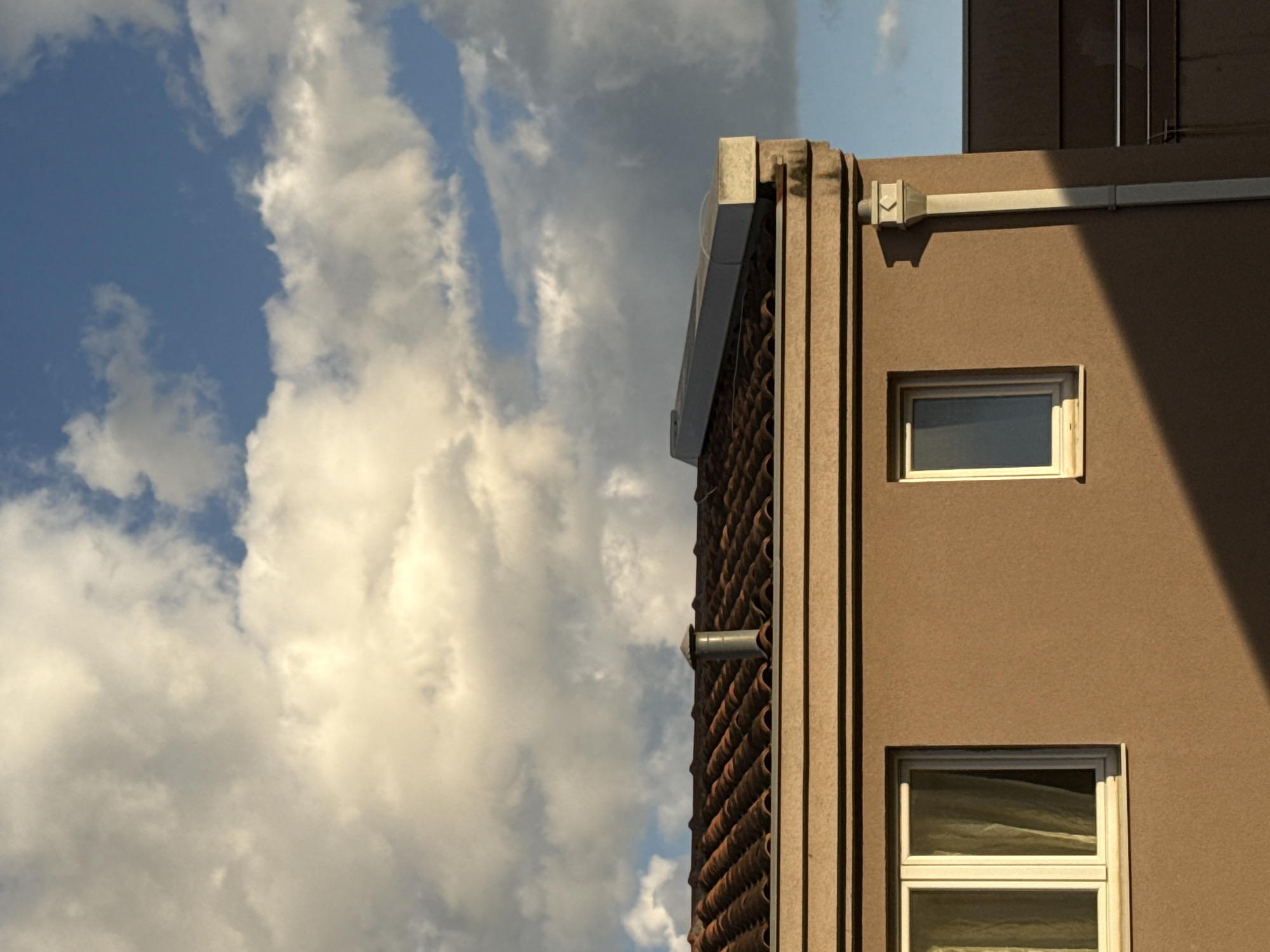 The image presents an upward view of a building facade against a dynamic sky. The building, located in Bar, Montenegro, is a warm brownish-beige color, with prominent vertical fluted architectural details on its corner.
On the right side of the building, two rectangular windows with white frames are visible; the top one is smaller than the bottom. A light-colored conduit runs horizontally across the upper part of this wall, leading to a square-shaped fixture near the corner. A distinct, sharp diagonal shadow cuts across the upper right section of the building and the top window, indicating strong directional sunlight.
The side of the building, visible behind the corner detail, features a textured surface composed of repetitive, reddish-brown, possibly terra cotta-colored, elements arranged in a patterned screen or grill. A small, round metal vent pipe extends from this textured section.
The sky occupies the left two-thirds of the frame, showcasing patches of clear blue interspersed with large, billowy white and light grey clouds. The lighting suggests either morning or late afternoon due to the long shadows. No people or activities are depicted, and no text is visible. The scene captures an architectural detail set against an expansive sky.