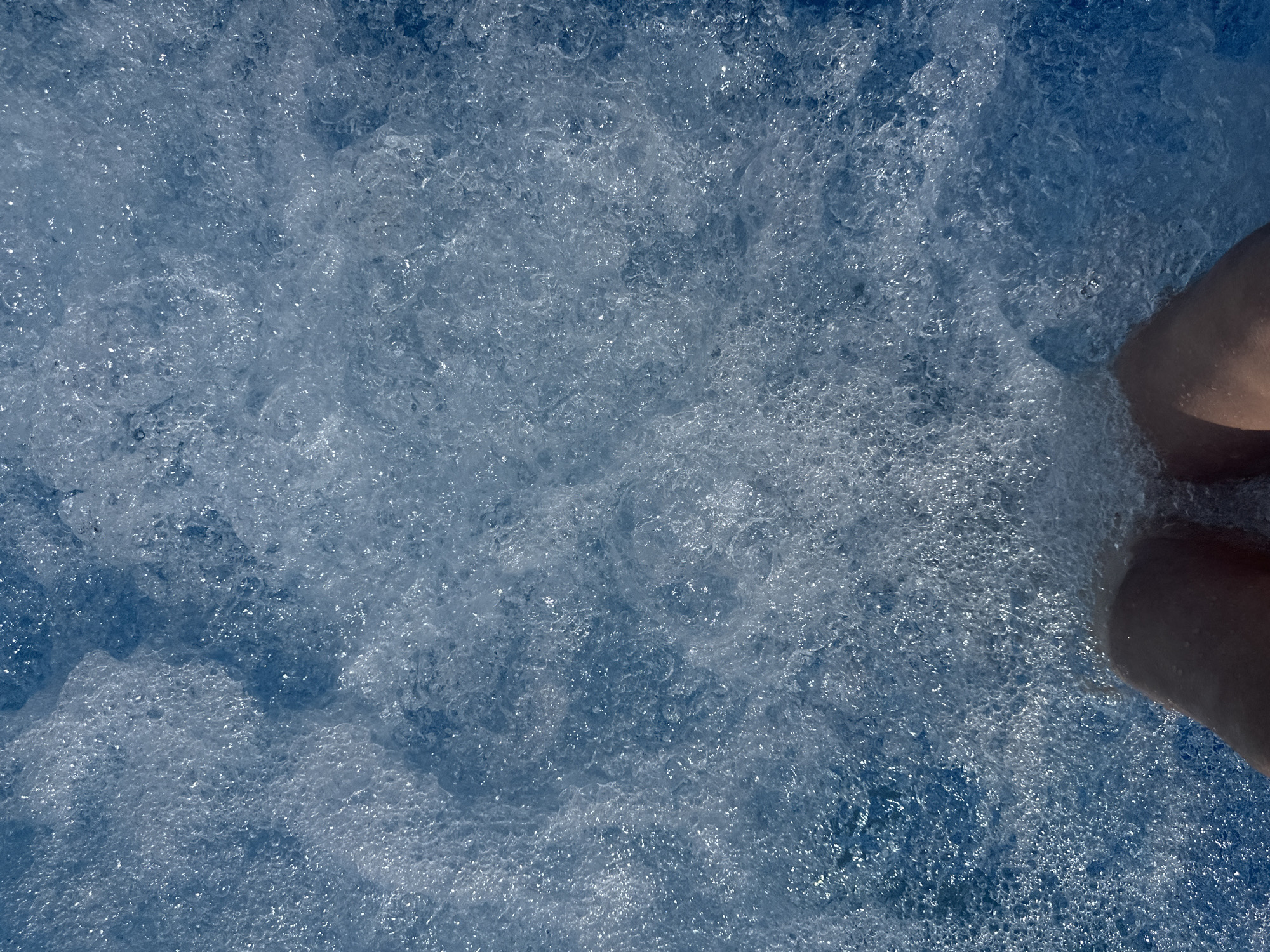 A close-up, overhead shot captures the frothy, blue water of a swimming pool or hot tub. The water is filled with countless small bubbles, creating a textured, opaque surface with highlights from the light reflecting off the water. Partially submerged at the bottom of the frame are two bare feet and lower legs, with skin appearing to be tanned. The scene is static and focuses solely on the water and the feet. The location in Даниловка (алтайский), Russia, is implied by the provided context but not visually evident in the image itself.