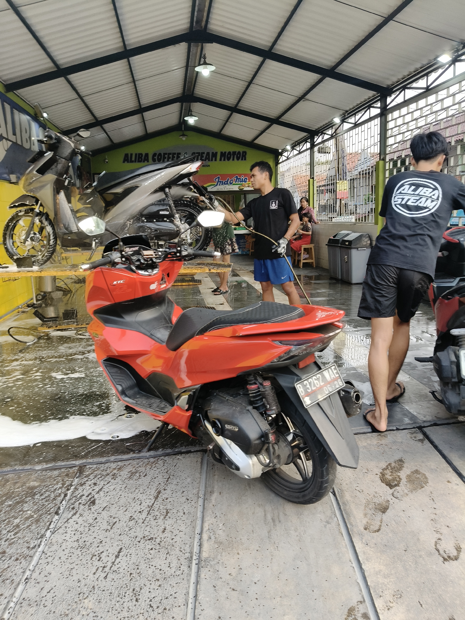 The image depicts a motorcycle washing service at "ALIBAB COFFEE STEAM MOTOR" located in South Tangerang, Indonesia. A bright orange scooter is positioned in the foreground, partially covered in soapy water. Behind it, a dark-colored motorcycle is on a yellow platform, undergoing washing. Two individuals are present; one, wearing blue shorts and a black t-shirt, is actively washing a motorcycle with a hose. Another person, wearing black shorts and a black t-shirt with a circular logo, stands nearby, facing away from the camera. The environment is an open-air wash bay with a metal roof structure, concrete flooring, and metal grates. The scene is consistent with a bustling local business catering to motorcycle maintenance and cleaning.
