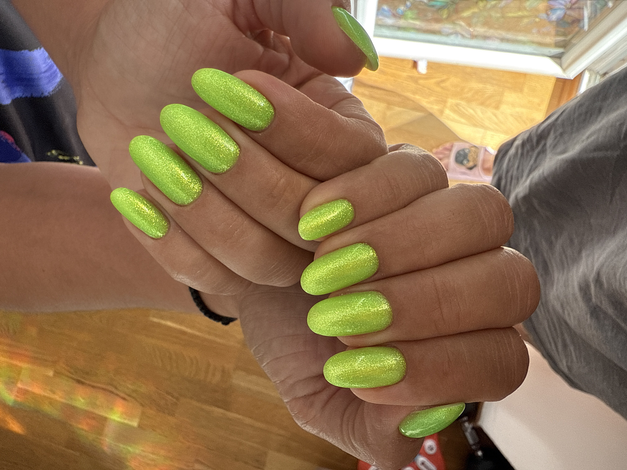 This media file features a close-up shot of a person's hands with bright, neon green, glittery nails. The nails are oval-shaped and uniformly painted with this eye-catching color. The hands are positioned in a way that displays multiple nails from different angles. One hand is held over the other, creating a layered effect.
In the background, there are hints of a domestic setting. A wooden floor is visible, with a faint rainbow-like reflection cast upon it, possibly from sunlight interacting with an object or window. A gray, textured fabric, perhaps a blanket or piece of clothing, is also present in the background, along with what appears to be the edge of a table or counter. Above, a window or glass surface shows a blurred reflection of what could be a colorful picture or scene.
The overall impression is one of personal grooming and style, with a focus on the vibrant nail color. The lighting suggests it might be daytime, given the natural light effects. There is no visible text in the image. The focus is entirely on the aesthetic of the nails and the immediate hand details.