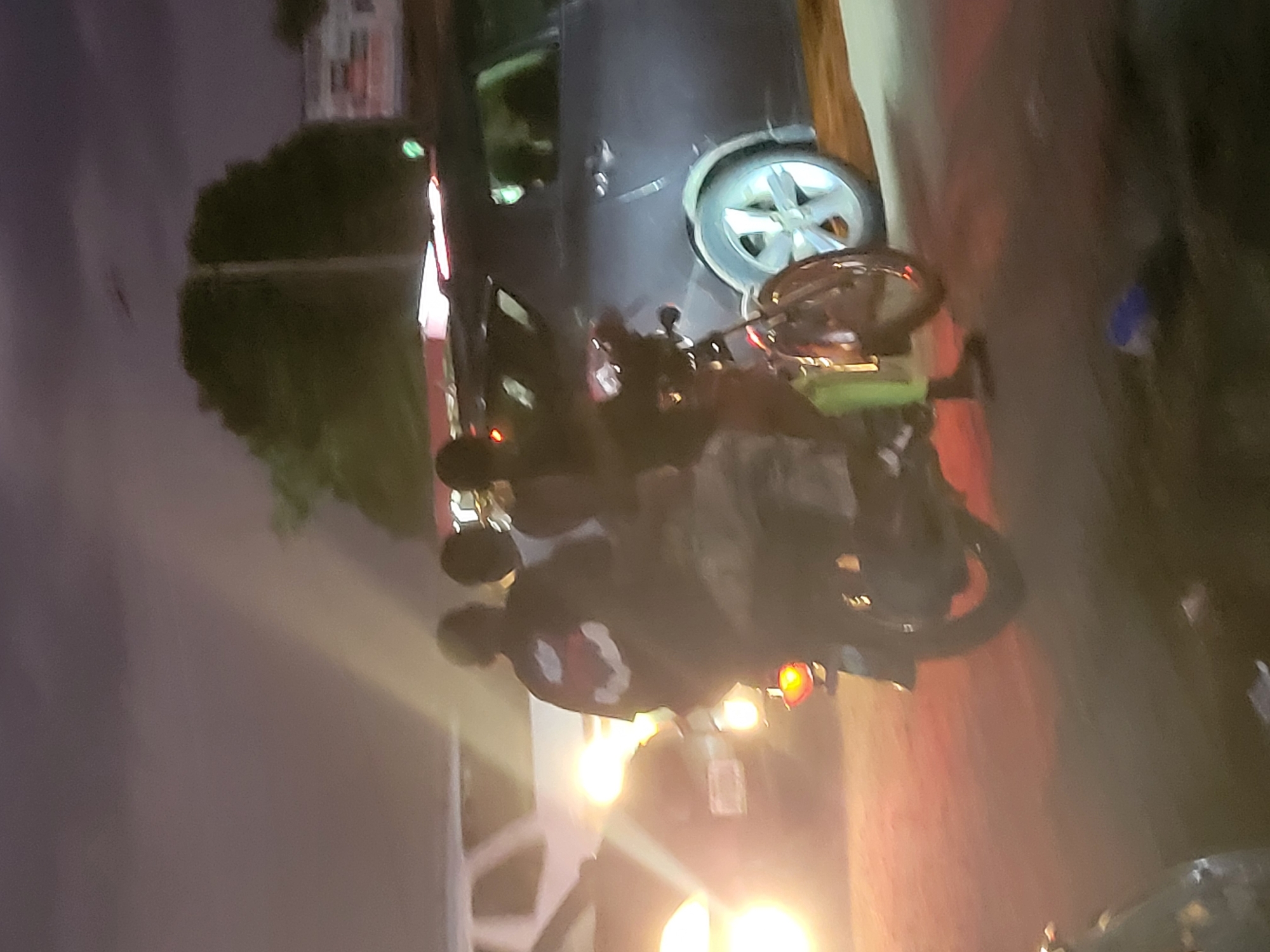 Three individuals are on a motorcycle at what appears to be a nighttime street scene in Kwamba, Nigeria. The motorcycle is dark-colored and has a visible rear taillight. The rider in the front is wearing a dark shirt with a red design on the back, and a lighter-colored garment is visible on the person behind them. The third person is partially obscured. They are stopped on a paved surface, with other vehicles and structures visible in the background, suggesting an urban or semi-urban setting. The lighting suggests it is evening or night.