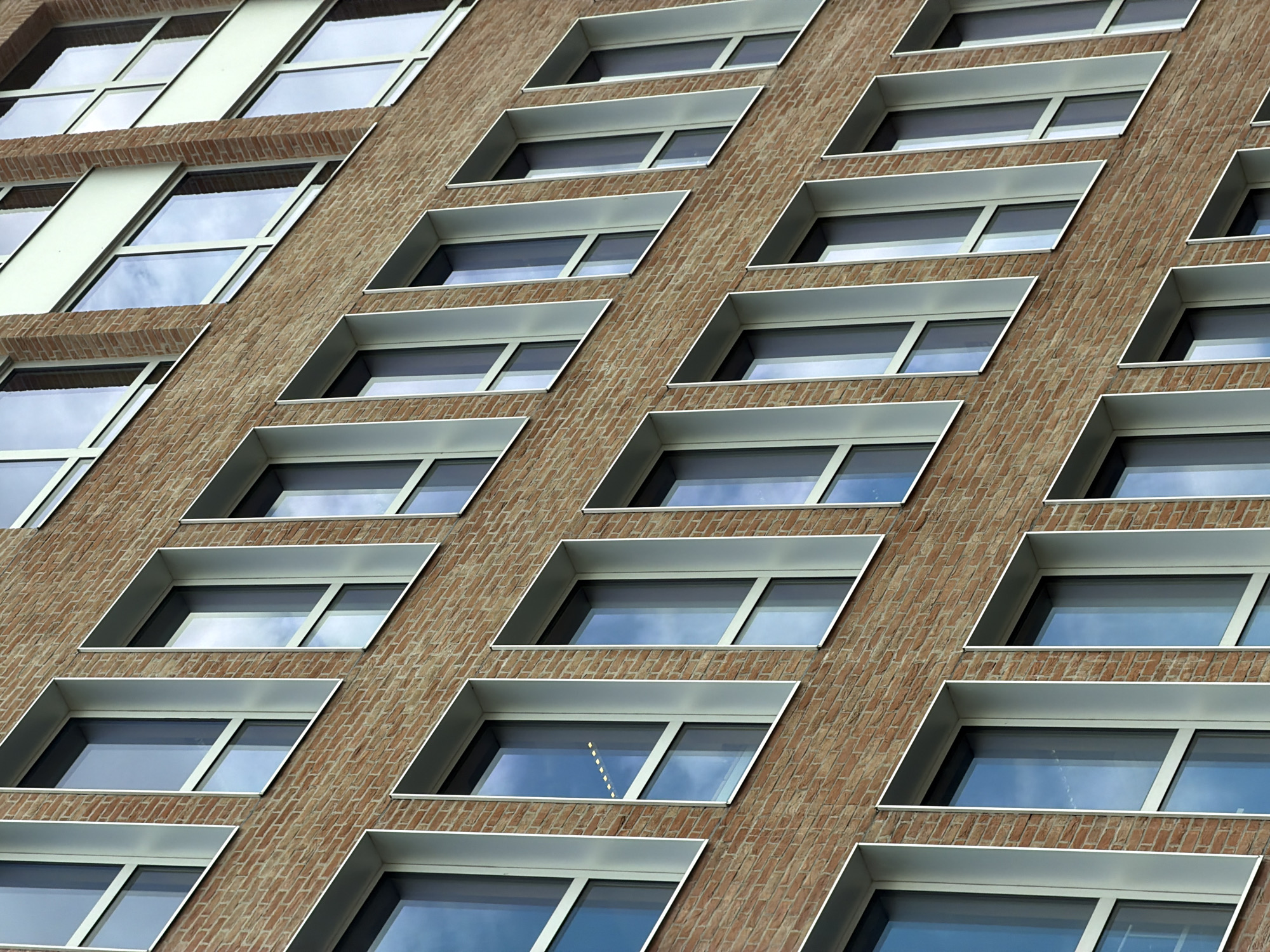 The image displays a close-up, diagonal view of a modern brick building's facade, characterized by its repetitive pattern of windows. The building appears to be located in Ivry-sur-Seine, France. The facade is constructed from warm-toned bricks laid in a regular pattern. Each window is set within a clean, angular, white frame that projects outwards from the brickwork. These frames create a distinctive, layered effect across the building's surface. The windows themselves are large and reflective, mirroring a cloudy sky, suggesting an overcast day. The building's architectural style is minimalist and geometric, with a strong emphasis on repeating modules. No people are visible in the frame. The scene is one of urban architecture, highlighting the structural and aesthetic design of the building. There is no visible text in the image.