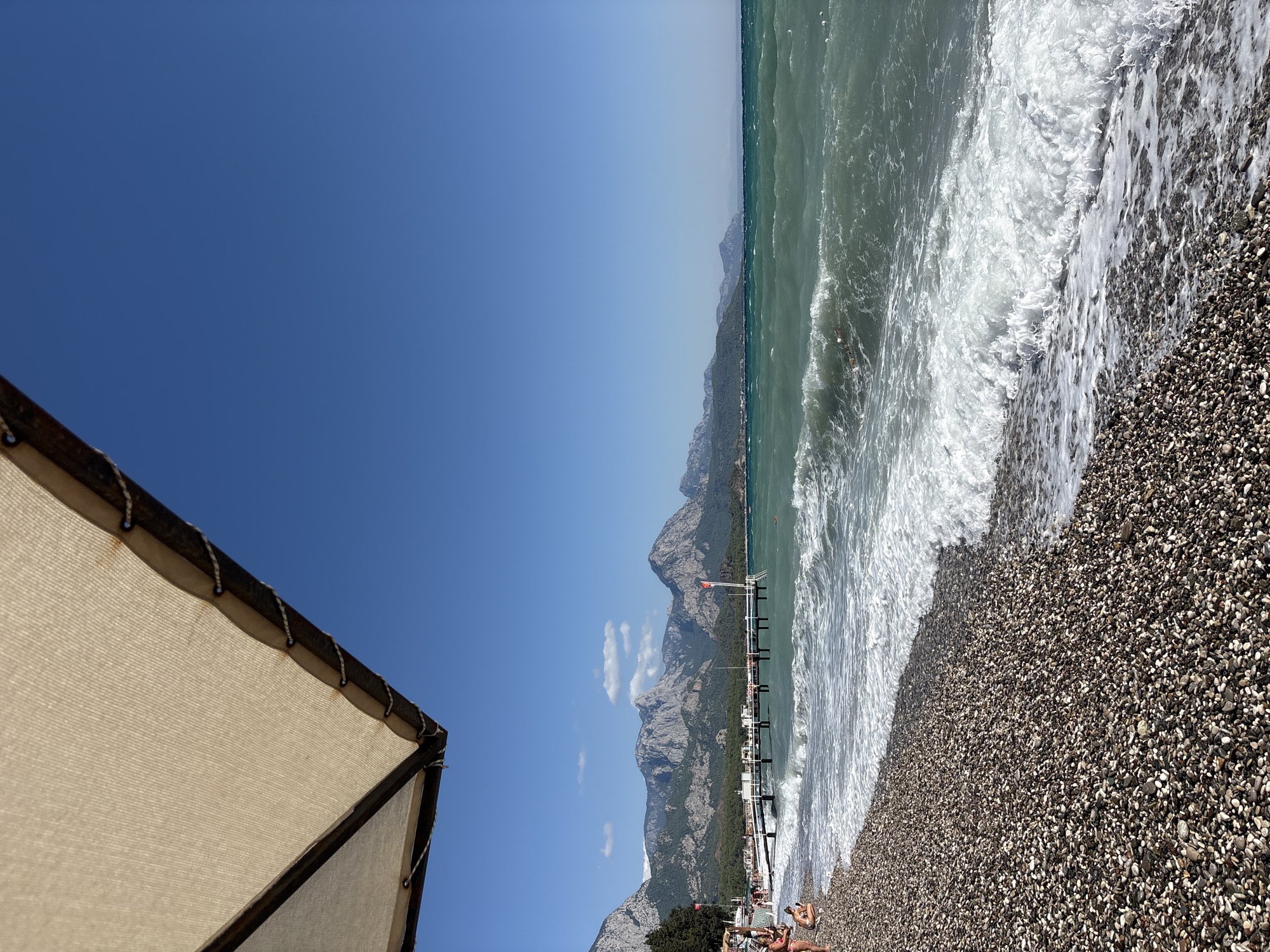A pebble beach in Kemer, Turkey, is bordered by a body of water under a clear blue sky. In the foreground, the pebbles are a mix of dark and light tones. Gentle waves break onto the shore, creating white foam. In the mid-ground, a long pier extends into the water, lined with white railings and some recreational facilities. Beyond the pier, a line of trees separates the beach from a range of rugged mountains that dominate the background. The mountains are covered in greenery, with rocky outcrops visible. On the left side of the image, a few people are visible on the beach, some sitting on the pebbles, and others wading near the water's edge. A section of a large, light-colored umbrella is visible in the upper left corner, providing shade. The overall scene depicts a sunny day at a coastal resort.