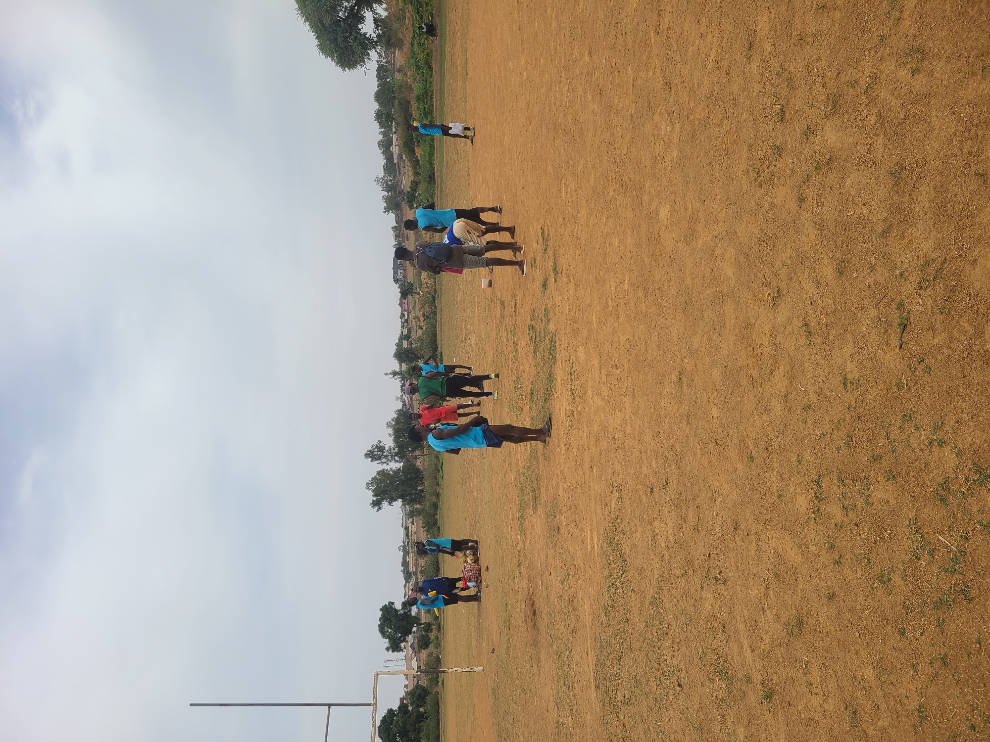 A wide shot captures a group of individuals on a field in Barikin Jirga Summa, Nigeria. The ground is dry and sparsely covered with green vegetation. A sports field with goalposts is visible. The people are wearing varied clothing, including blue, red, and green shirts. They appear to be exercising or training. The sky is overcast, with patches of white clouds. Trees and shrubs line the edge of the field.