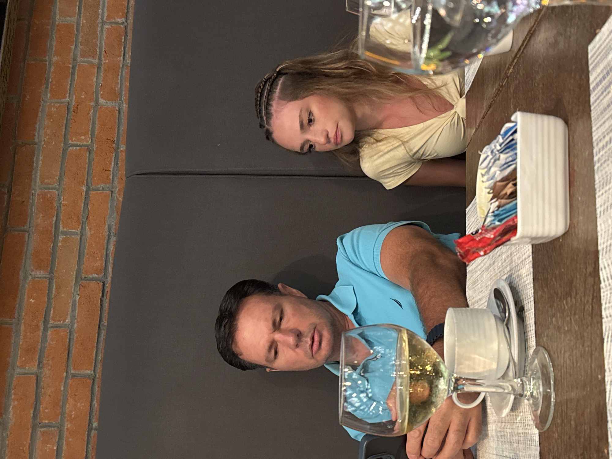 A man and a young woman are seated next to each other at a table in what appears to be a restaurant, possibly in Playa del Carmen, Mexico. The restaurant's interior features a brick wall and a padded gray booth. The table is set with a beige table runner. In front of the man are a smartphone, a wine glass filled with a light-colored liquid, and a cup and saucer. The woman has a glass of a light-colored liquid in front of her and a dispenser containing sugar packets. The man is wearing a blue polo shirt. The woman is wearing a yellow top. They both appear to be looking at the table or possibly at something on the man's phone.