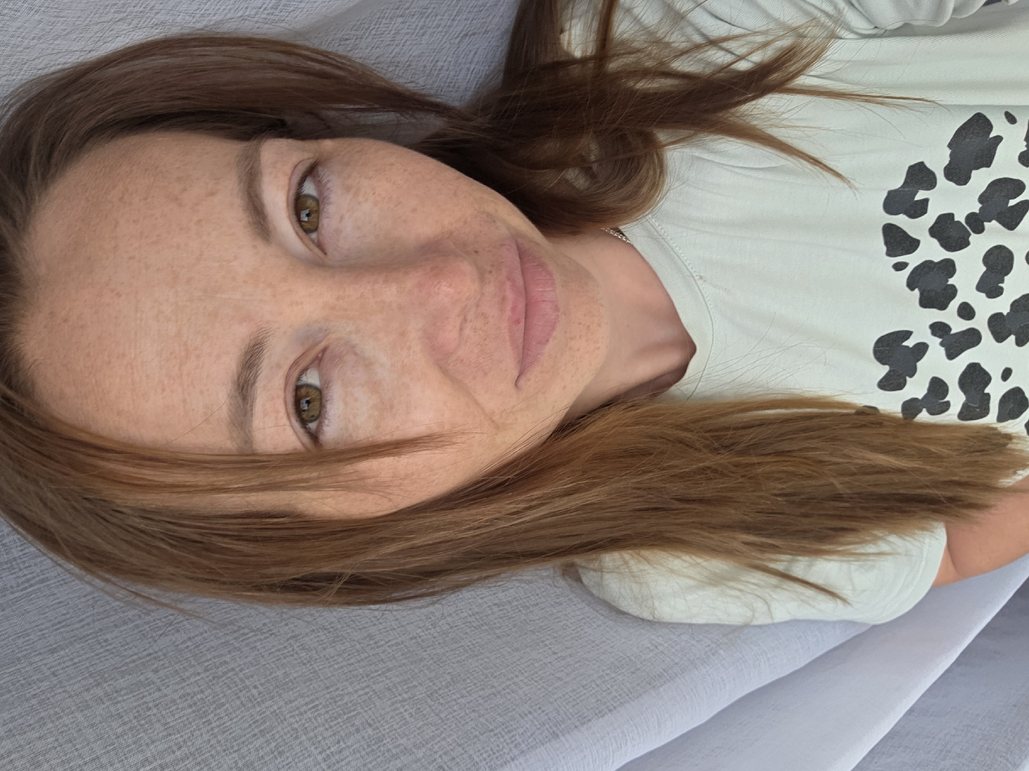 A woman with auburn hair and freckled skin looks directly at the camera. She wears a light-colored shirt with a black spotted print. Her background consists of white fabric. The location is Unknown, Unknown.