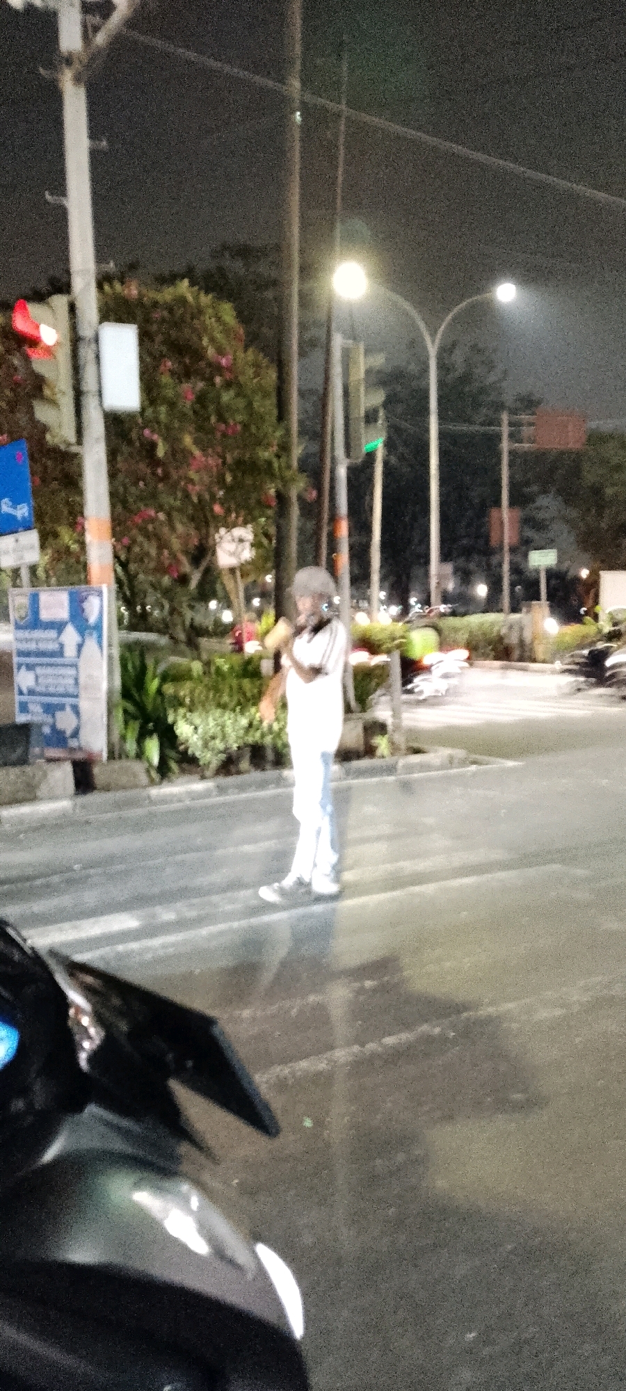 The image captures a nighttime scene at a city intersection in Depok, Indonesia. A pedestrian, appearing to be an adult male wearing a white long-sleeved shirt and light-colored pants, stands on the crosswalk. He is facing towards the right of the frame and appears to be holding something to his mouth, possibly a cigarette or a mask, while looking away. The environment is illuminated by streetlights, with a traffic light showing green in the background on the right side of the frame. Trees and foliage are visible behind the pedestrian, and traffic signs are present on the left. The foreground shows the front of a dark-colored motorcycle, with its front fender and handlebars partially visible, suggesting the photo was taken from a parked motorcycle. The road surface is asphalt, and there are white stripes indicating the pedestrian crossing. The overall image has a grainy quality due to the low light conditions.