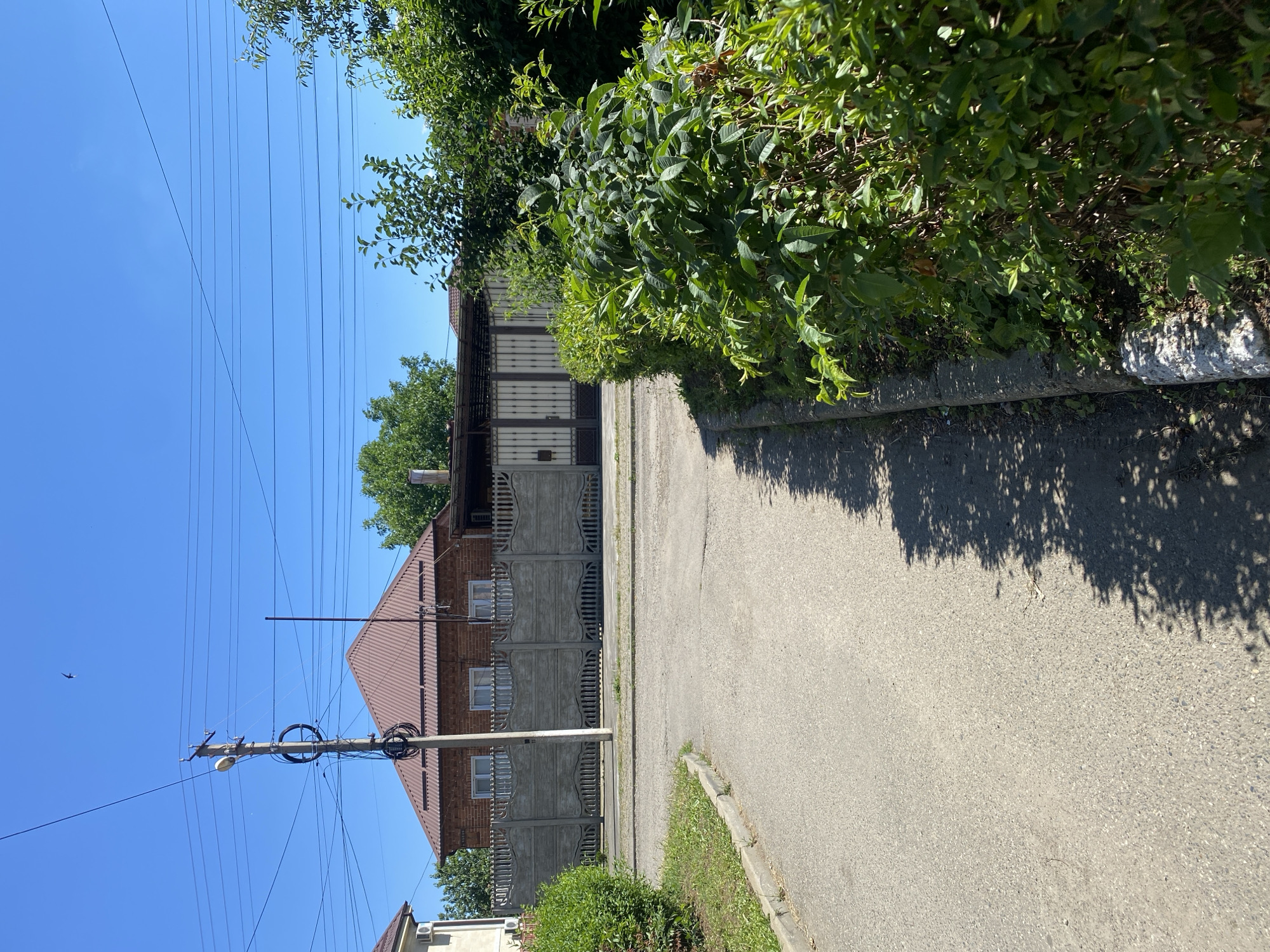 A paved road curves into the foreground, leading towards a long, decorative concrete fence with arched details. Behind the fence stands a two-story brick house with a reddish-brown tiled roof. A tall utility pole with numerous wires and a light fixture is situated in front of the house. To the right of the road, a dense green hedge lines the curb, casting shadows onto the pavement. Beyond the fence, a wide, light-colored metal gate is visible. The sky is a clear, bright blue. The scene appears to be a residential area in the city of Zapadny, Russia.