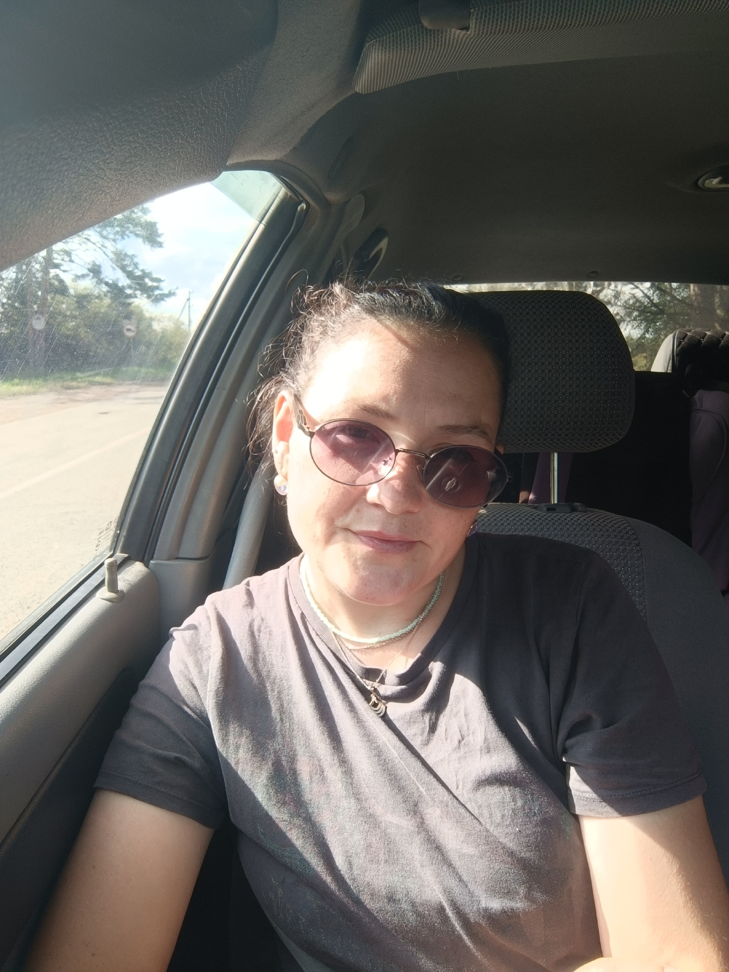 A medium shot photo shows a woman in the passenger seat of a car, looking at the camera. She has dark hair pulled back, is wearing sunglasses with rounded lenses and a pearl earring. She is wearing a dark gray t-shirt and a light green beaded necklace with a pendant. Sunlight streams in through the window on the left, casting shadows on her face and shirt. The interior of the car is visible behind her, including the headrest and part of the seat. Outside the window, a road and trees can be seen under a bright sky. The exact location in Foki, Russia is not specified in the image.