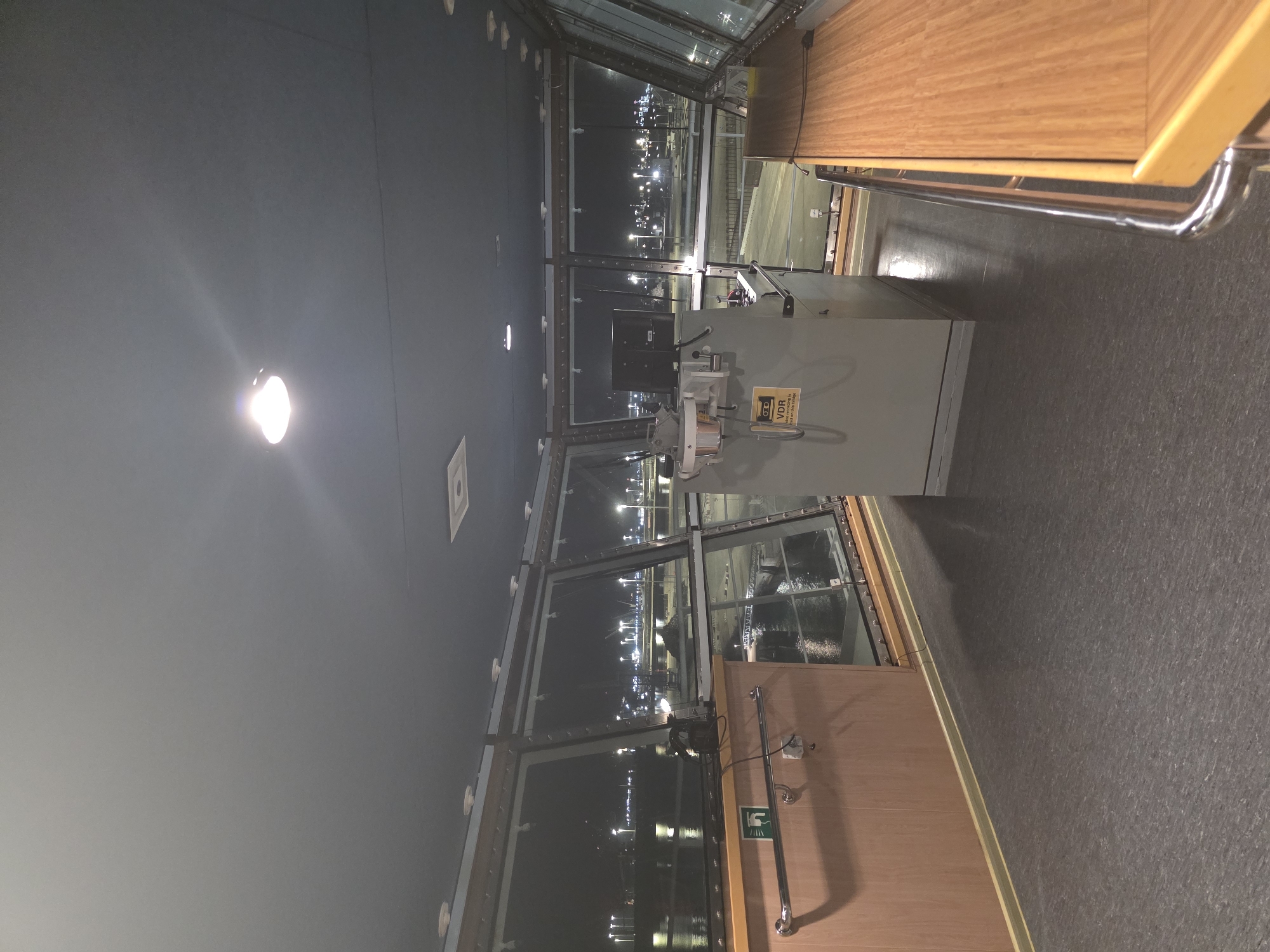The image shows the interior of a ship's bridge at night, overlooking a brightly lit port. The bridge is equipped with large windows that offer a panoramic view of the port facilities, including numerous lights, cranes, and water. Inside the bridge, there is a console with electronic equipment and a computer monitor. A railing runs along the side, and wooden cabinetry is visible. The floor is dark gray and appears to be made of linoleum. There are no people visible in the image. The overall impression is one of a functional, operational environment within a vessel docked at a port.
