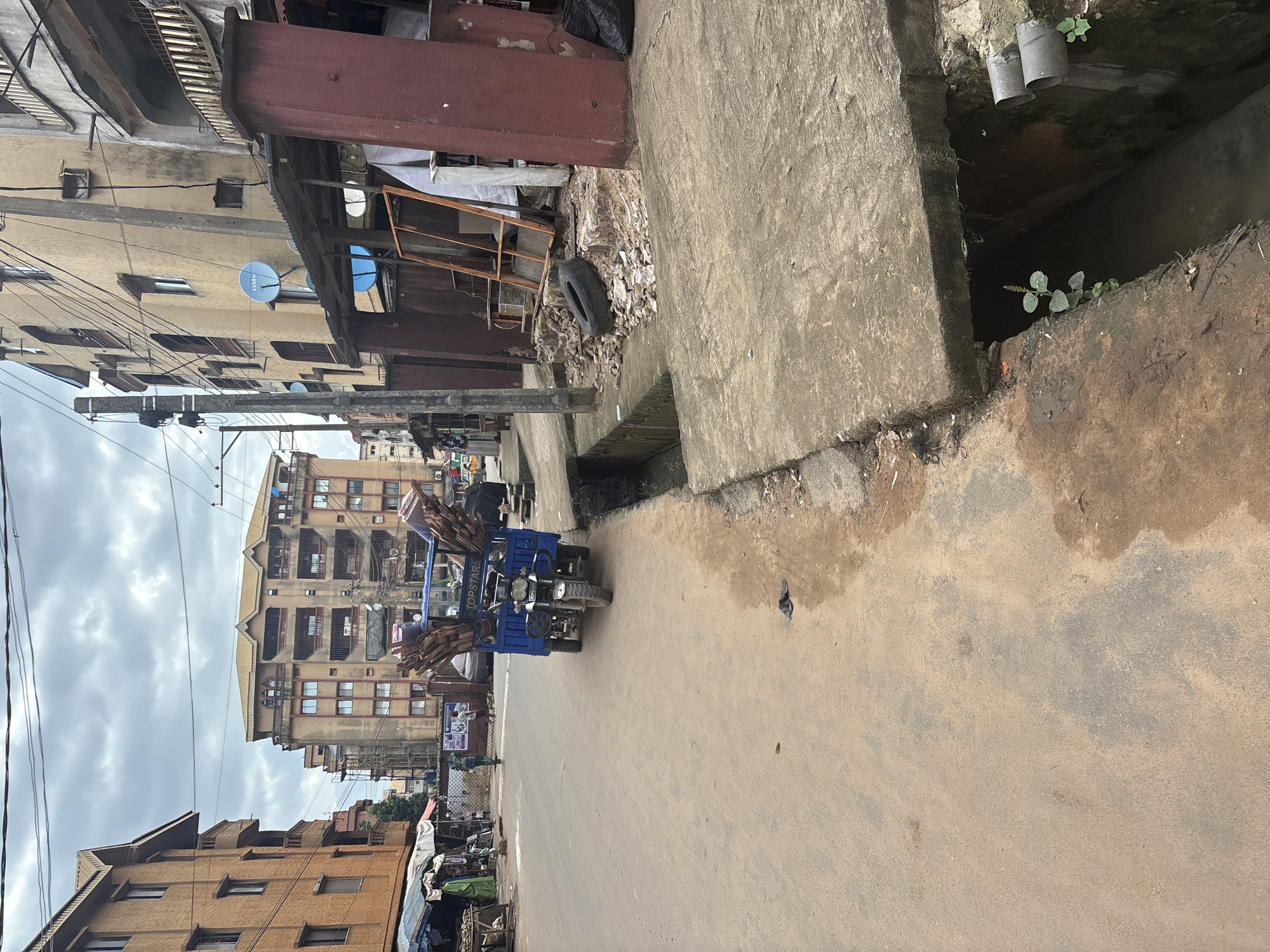 This is a street-level, eye-level, medium shot of a street in Onitsha, Nigeria. The street is paved with a reddish-brown earth-like material, with patches of grey asphalt. On the right side of the street, there is a concrete gutter and a sidewalk. A blue motorized tricycle, commonly known as a "keke," is parked in the middle of the road, loaded with lumber. The tricycle has the word "TOPSTAR" written on its front.
Several buildings are visible in the background. On the left, there are multi-story buildings with balconies and cream-colored walls. On the right, a taller building with a yellowish facade and several satellite dishes on its exterior dominates the view. In front of this building, there is a covered market stall with various items displayed, and a section with debris, a tire, and wooden frames.
Two individuals are visible. One person, wearing a green garment, is walking away from the camera on the left side of the street, near the market stalls. Another person, appearing to be a child, is standing further down the street near the tricycle.
The sky is overcast with grey clouds. The overall impression is of a busy urban environment with a mix of residential and commercial structures.