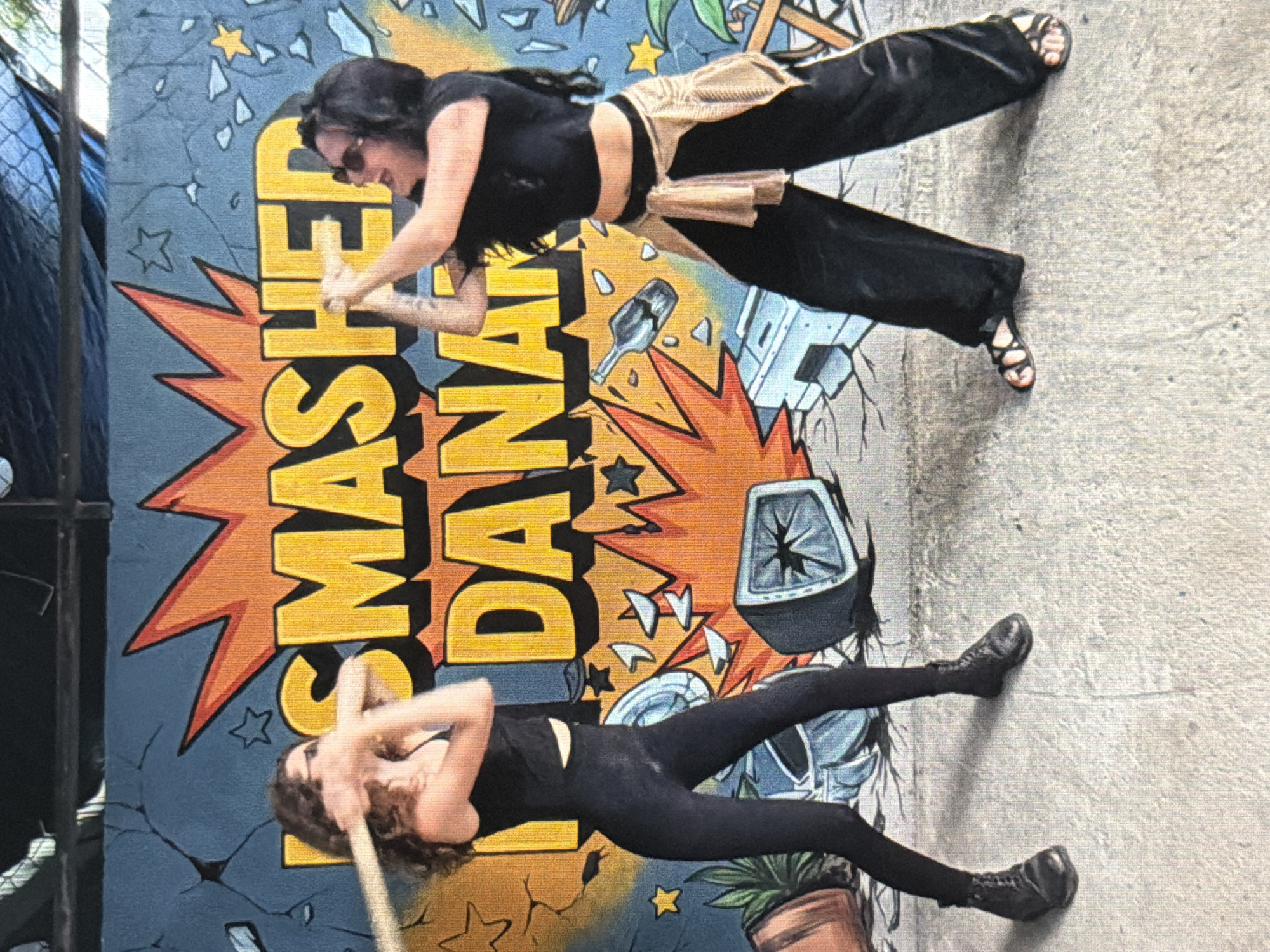 A medium, eye-level shot shows two women posing in front of a mural. The mural depicts an explosion with the words "Smashed Banana" in yellow. One woman, positioned above, wears black pants and a black crop top with a brown sash. She has black hair and wears sunglasses, and she's captured mid-action, swinging her arms. The second woman, positioned below, wears all black, including leggings and boots, and is swinging a wooden stick. The background shows a concrete ground on the right and a fence on the left. The location is unspecified; there is no indication of Đà Nẵng, Vietnam.