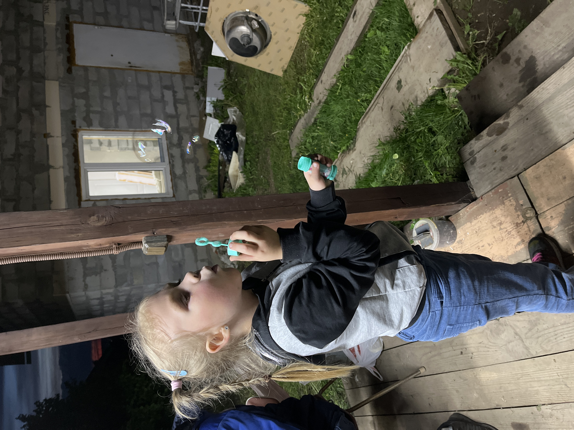 A young, light-haired girl wearing a black and grey jacket and blue corduroy pants is standing on a wooden surface, possibly a porch or deck. She is blowing bubbles with a green wand, and a few bubbles are visible in the air near her face. Her hair is styled in pigtails with blue and pink hair accessories.
The environment consists of a building with concrete block walls, a window, and a white door. There is a piece of furniture or construction material near the wall. Green grass and foliage are visible. The location is Chaikovsky, Russia.