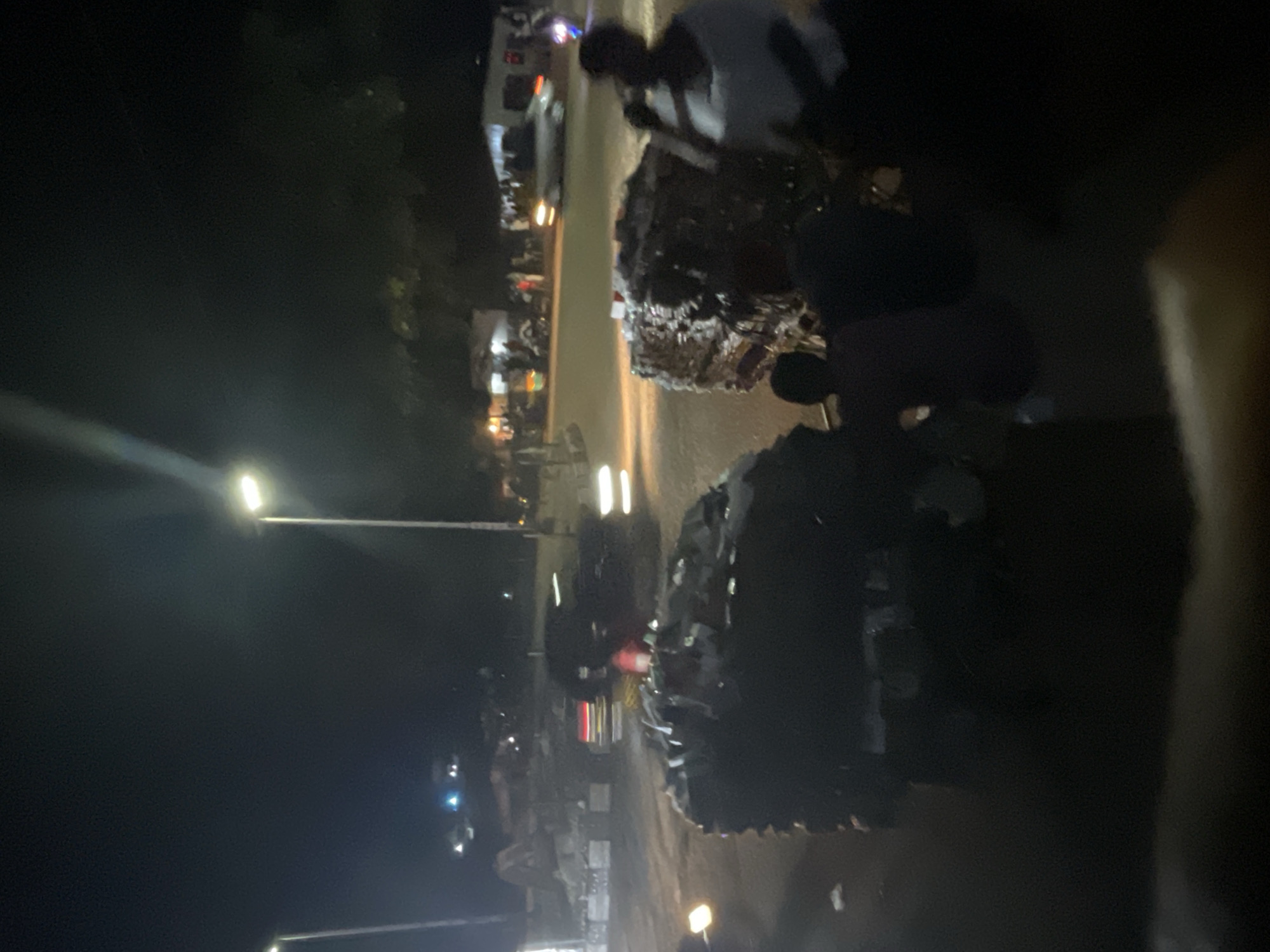 At night, on an outdoor paved surface, people are gathered near large piles of compressed materials, possibly recycled goods. The scene is illuminated by streetlights and vehicle lights, creating a somewhat blurry and dark atmosphere. In the background, vehicles are moving on the road, and structures that appear to be buildings or stalls are visible. The people are mostly silhouetted against the lights, with some individuals appearing to be interacting or working with the piles. The overall impression is of a busy area at night, likely a market or a recycling depot.