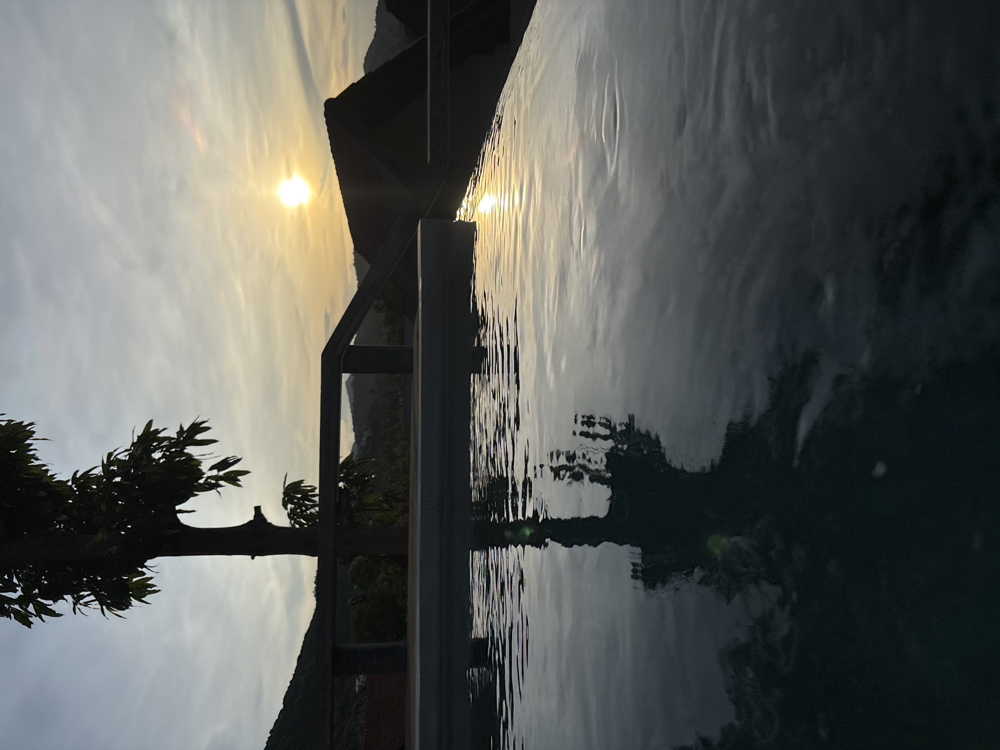 The image depicts an outdoor swimming pool at sunset. A silhouetted tree stands to the left of the frame, its reflection visible in the rippling water of the pool. The pool's edge is in the foreground, with the water surface taking up the lower half of the image. Beyond the pool, a wooden railing separates the pool area from a landscape of hills and rooftops. The sun is setting in the distance, casting a warm glow across the sky, which is partially obscured by clouds. The overall atmosphere is tranquil and serene.