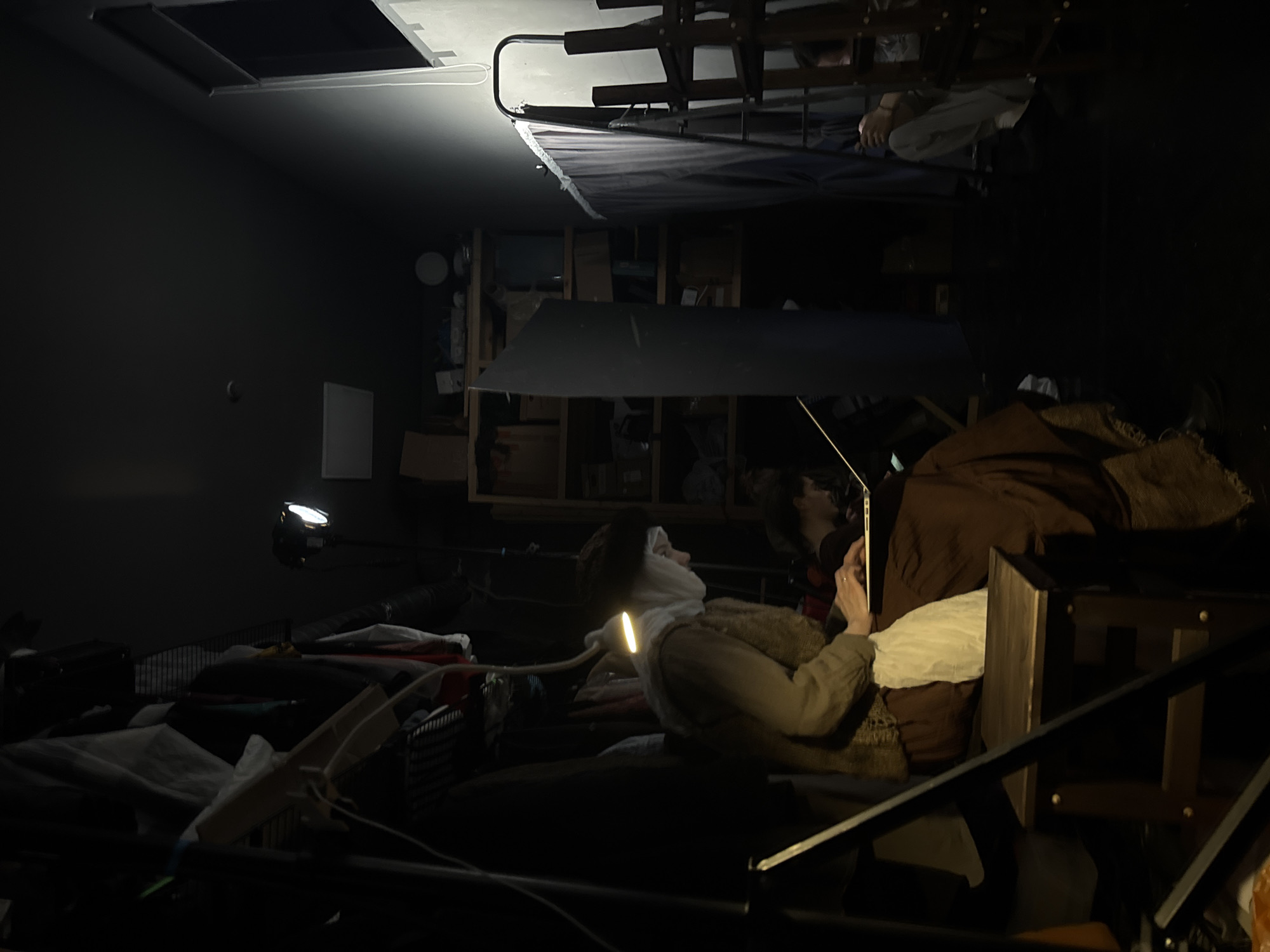 The image depicts the interior of a cluttered room with various objects and two individuals. One person is lying down, possibly on a bed, with a light illuminating their face and torso. They are using a laptop. The second person is also nearby, appearing to be interacting with the person on the bed. The room is filled with boxes, shelving units, and other miscellaneous items, creating a sense of disarray. A bunk bed is situated in the background. The location in Moscow, Russia is not discernible from the image.