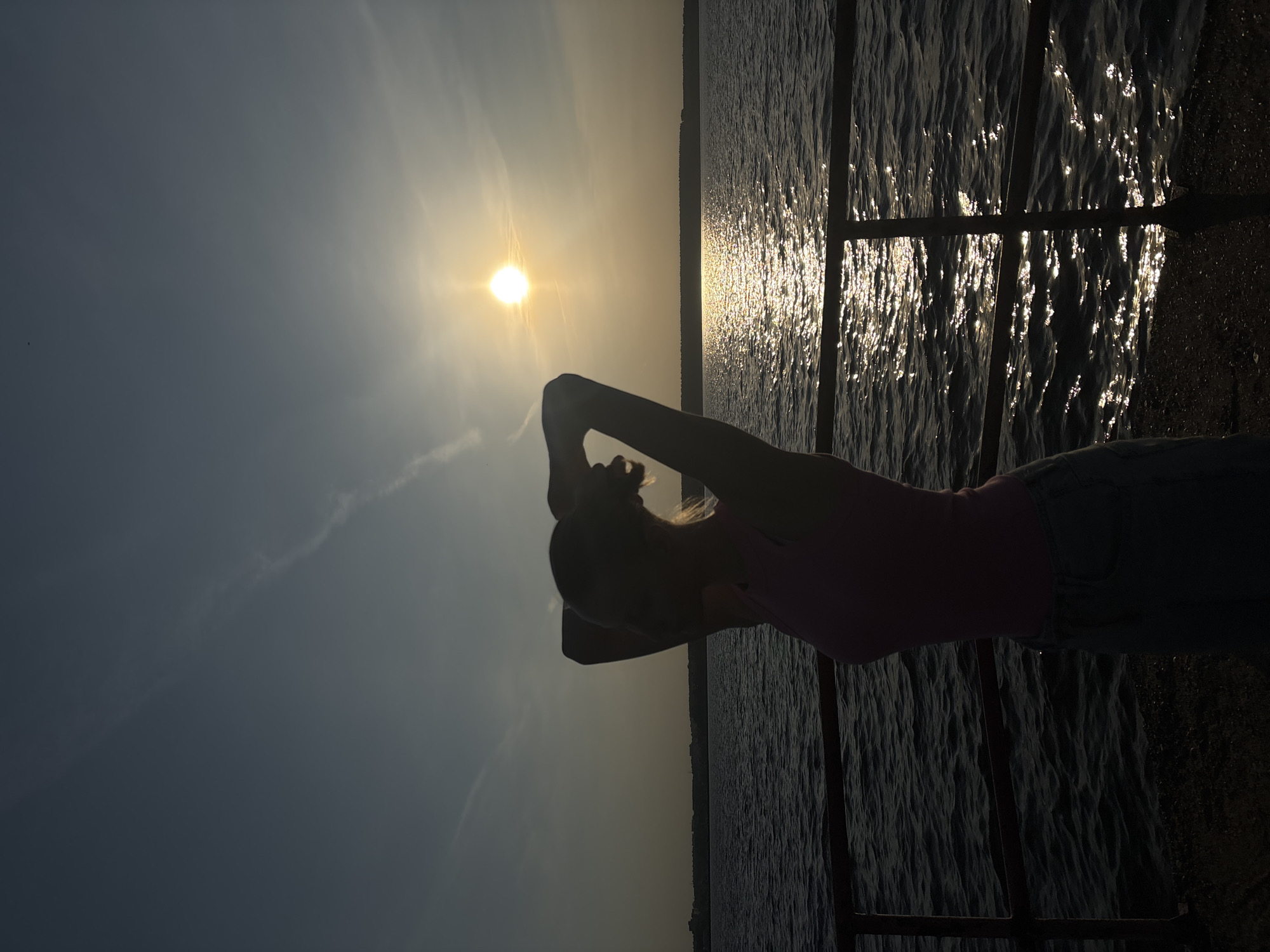 Here is a detailed description of the image:
**Scene Overview:**
The image captures a woman standing by a railing near a body of water, likely the Kama River, with the sun setting in the background. The scene is likely in or near Perm, Russia. The image is strongly backlit, rendering the woman in silhouette.
**People:**
* **Woman:** A single woman is positioned in the foreground, facing the water with her arms raised above her head. Her clothing is visible enough to discern a pink top and jeans.
**Objects:**
* **Water:** The Kama River, reflecting sunlight in a shimmering pattern.
* **Railing:** A metal railing is positioned in front of the woman, indicating a waterfront location.
* **Sun:** The setting sun is a prominent feature, creating a strong backlight that silhouettes the subject.
* **Sky:** The sky is visible above the sun, with some cloud formations.
**Environment:**
* **Location:** A waterfront area in Perm, Russia. The location could be on the Kama River embankment.
* **Weather/Time:** The photo was taken during sunset, with clear sky conditions, the time is in the evening.
* **Lighting:** The image is strongly backlit by the setting sun, creating silhouettes.
**Actions/Interactions:**
* The woman is standing by the railing, presumably watching the sunset. Her raised arms suggest a sense of freedom or enjoyment.
**Additional details**
* The water surface displays bright reflections of the sun, indicating calm conditions and clean water.
* The skyline across the river appears relatively flat.
* The image has been taken from a level slightly lower than the horizon.
* The woman is facing the river.
* The railing posts are metal pipes or bars with horizontal elements crossing them.
* A haze is present on the horizon.
* A contrail created by an aircraft is evident in the sky near the sun.
* In conclusion, the image captures a woman enjoying the sunset near the Kama River in Perm, Russia.