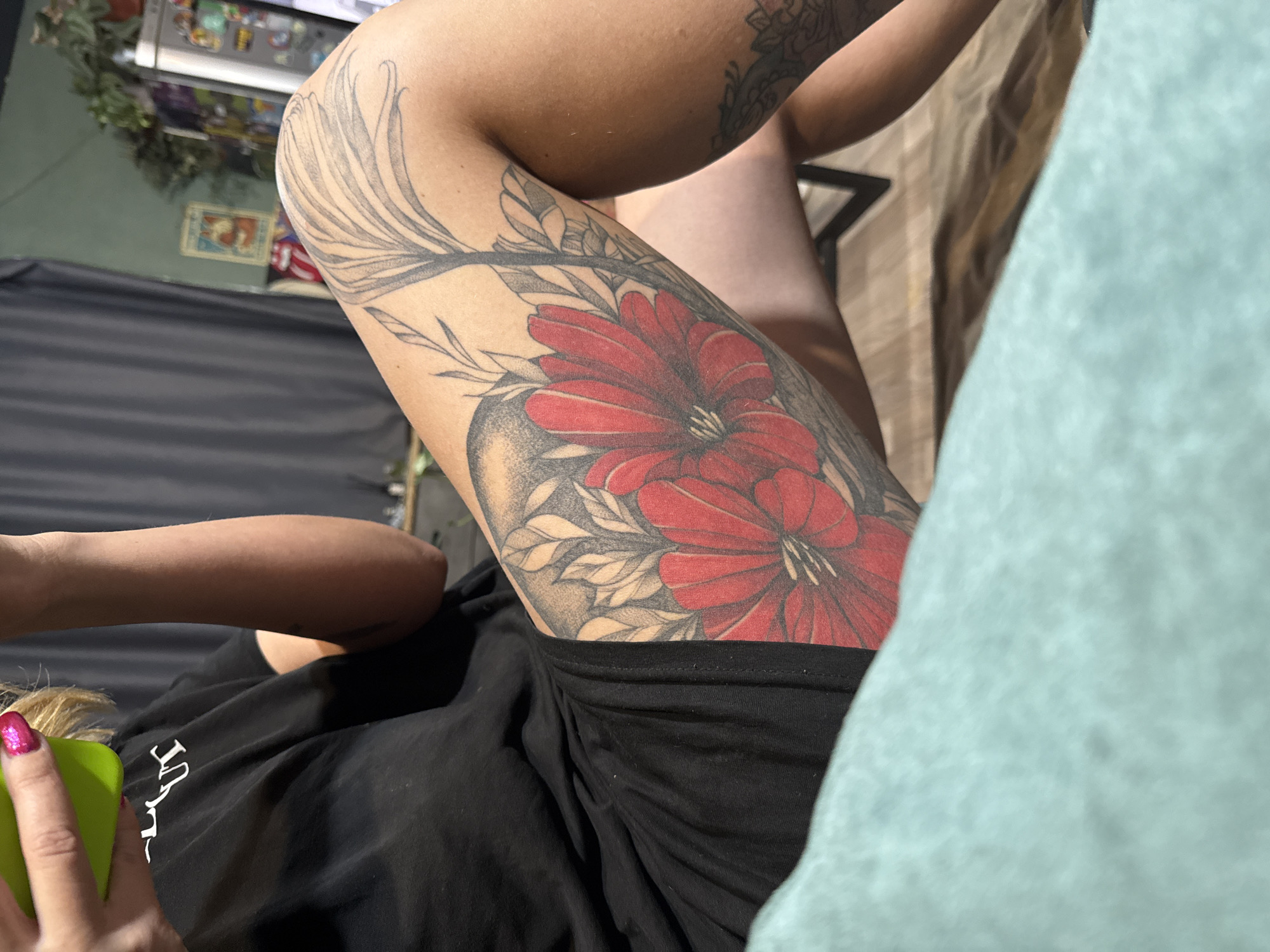 The image shows a person with tattoos on their arm and leg. The leg tattoo features red flowers and leaves, while the arm tattoo has a more intricate, pattern-based design. The person is wearing a black top and holding a green object. A light blue object is in the lower right. The background is somewhat blurred and includes furniture, and a wall. The location is not discernible from the image.