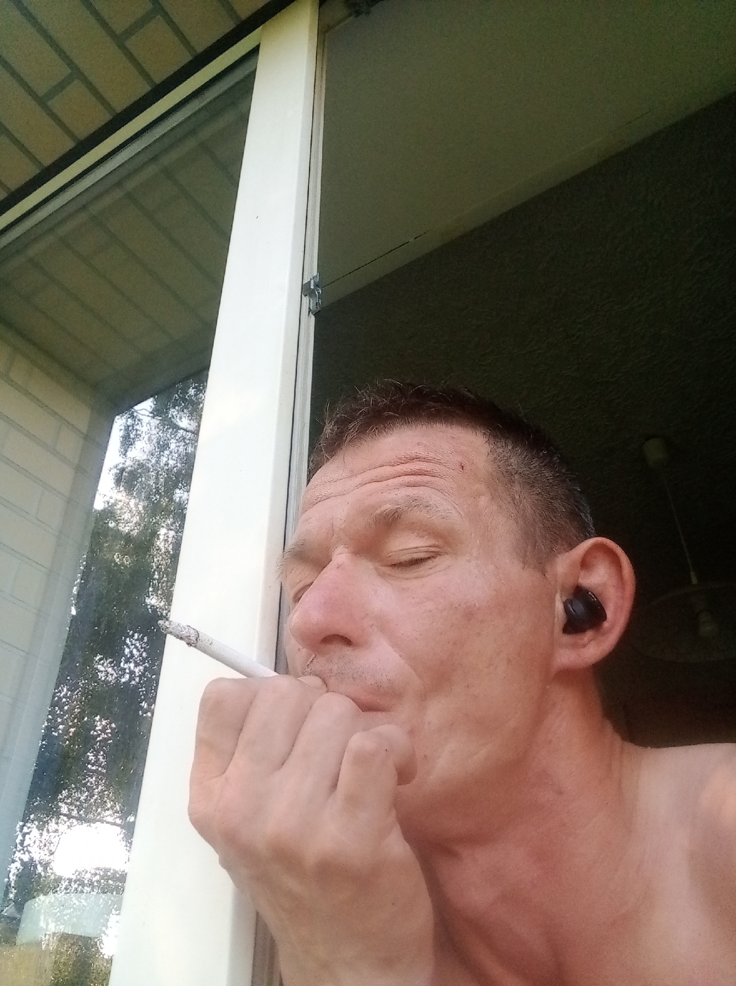A medium close-up, eye-level shot shows a shirtless man with short, dark hair and closed eyes, looking down and to his left. He is leaning his head against a white window frame. His right hand is raised to his face, with his thumb and index finger holding a lit cigarette, which he is about to bring to his mouth. He is wearing a black earbud in his right ear. Outside the window, trees and foliage are visible, suggesting an outdoor setting. The man's skin is weathered, and he has wrinkles on his forehead and around his eyes. The background consists of a dark interior space, likely a room, with a ceiling fan visible in the upper right corner. The man is indoors, looking out of a window. The image is not in Tapa, Estonia.