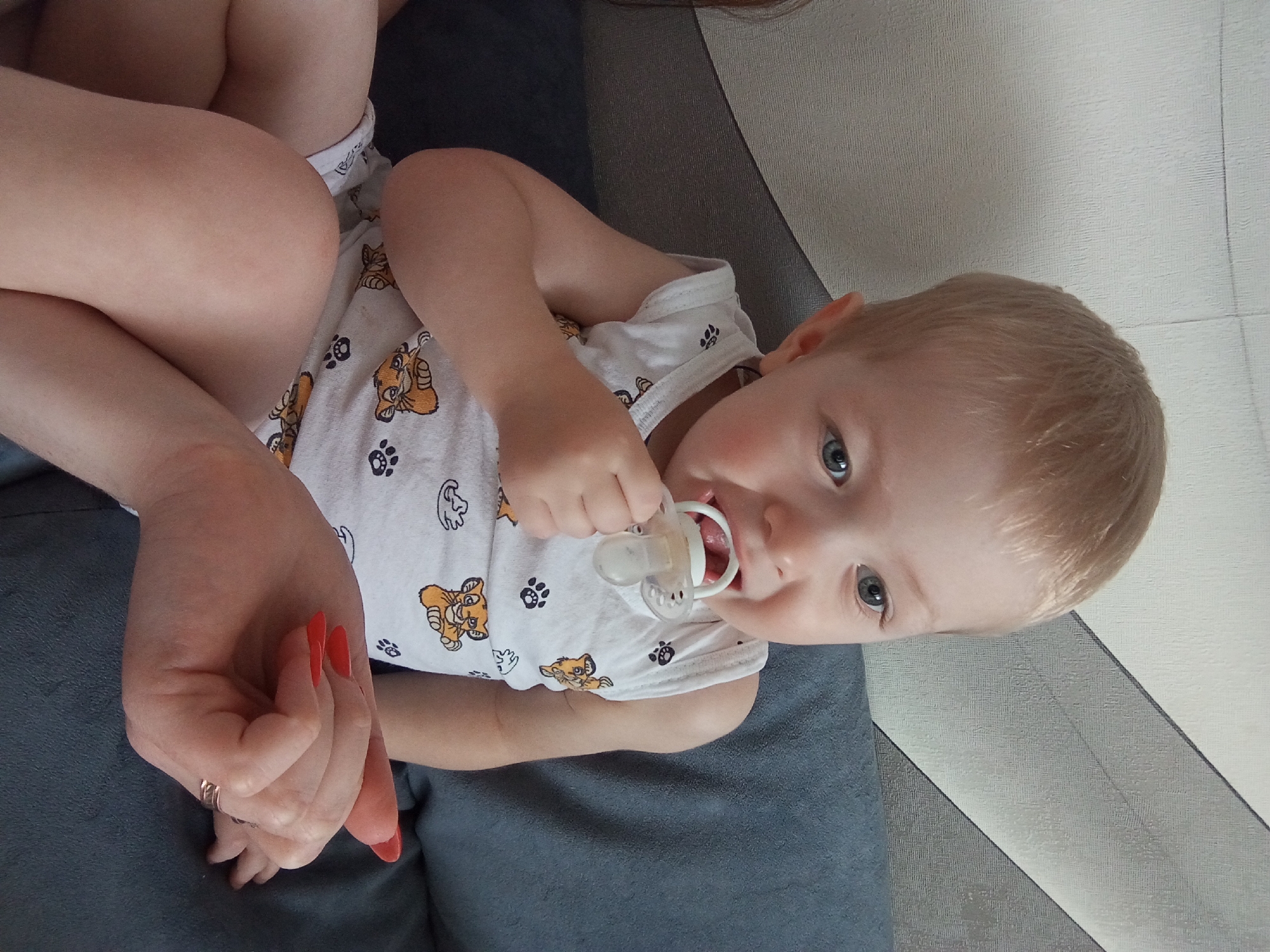 A close-up, slightly rotated image depicts a young child with blond hair and blue eyes, wearing a white onesie with Simba (from "The Lion King") and paw print patterns, with a pacifier in its mouth. The child is resting on a blue-gray upholstered surface. Part of an adult's hand with red nail polish and gold rings is visible to the left of the child. The background includes a plain white wall. The image location is Yurgamysh, Russia.