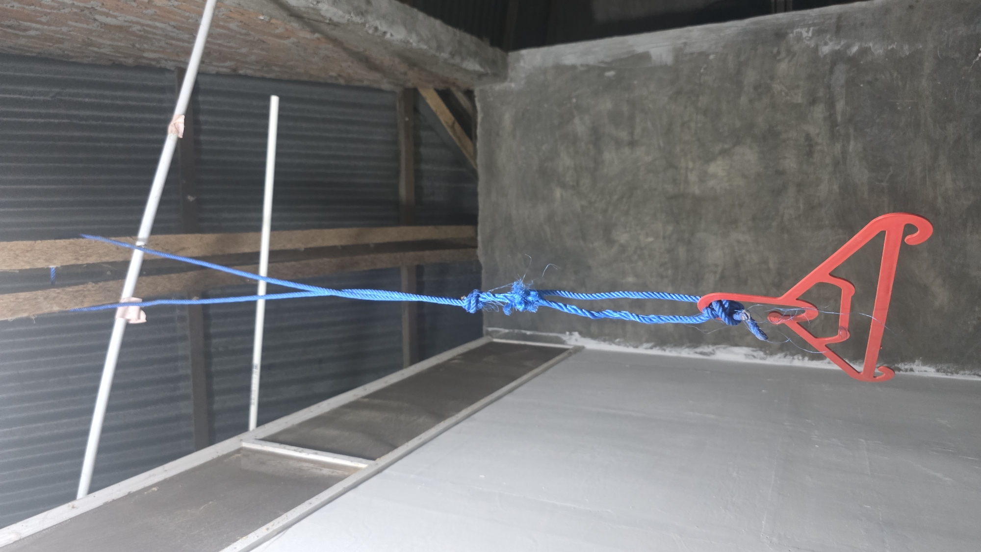 The image shows a makeshift clothesline inside a building in Lhokseumawe, Indonesia. A bright blue rope is strung between a white PVC pipe and a red plastic hanger. The rope is knotted in the middle. The background includes corrugated metal siding, wooden support beams, and a rough concrete wall. The floor appears to be painted grey.