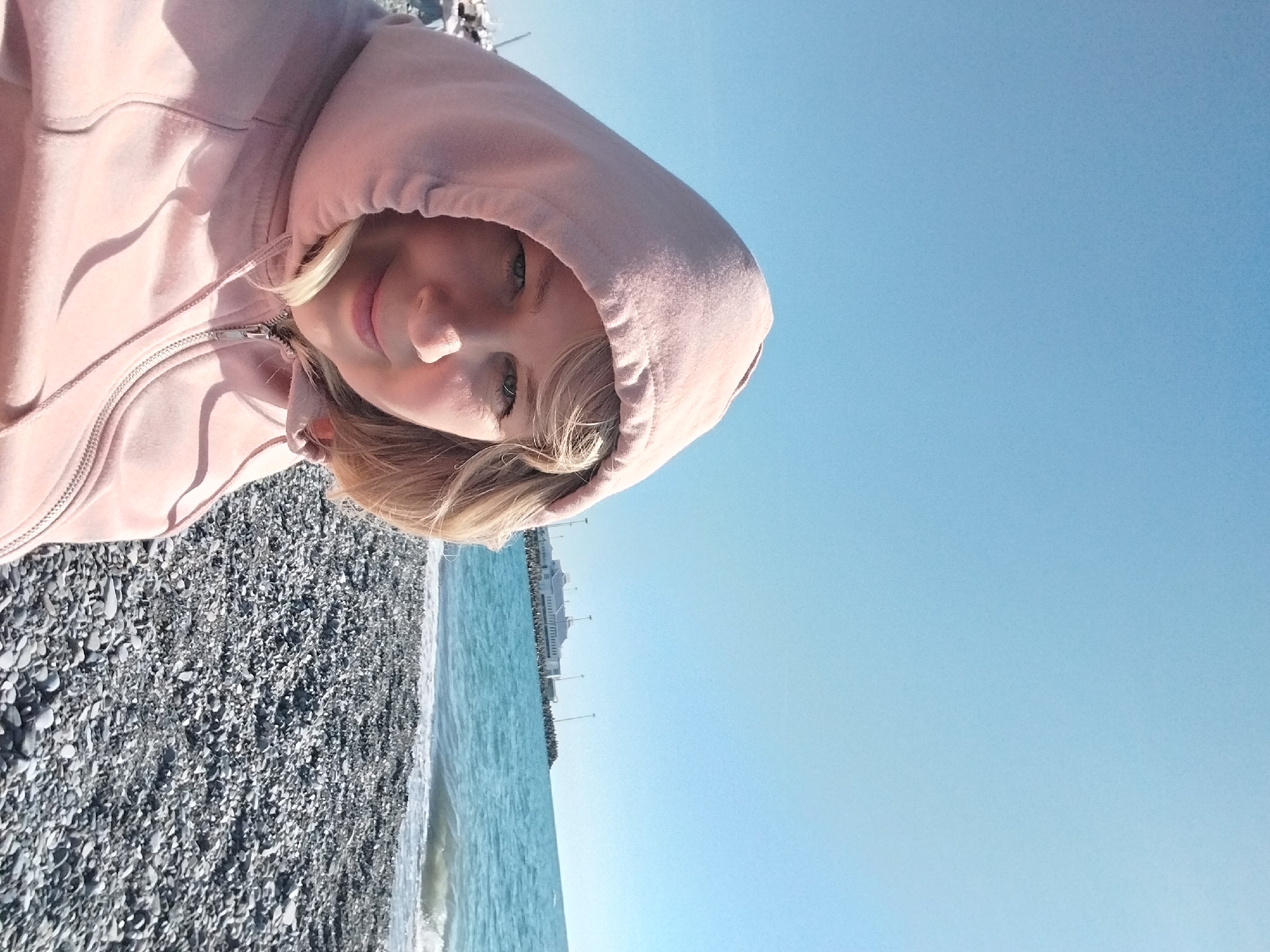 The image displays a close-up portrait of a young individual on a sunny pebble beach, rotated 90 degrees counter-clockwise from its likely original orientation. The subject, who appears to be a young female, is prominently featured in the upper left section of the frame (when rotated correctly). She is wearing a light pink hooded sweatshirt, with the hood pulled up around her head, framing her face. Her light blonde hair is visible, and she looks directly at the camera with blue eyes and a soft, contented smile. A silver zipper is visible on her sweatshirt.
Behind and to the side of the person, the scene transitions to a clear, bright environment. The extensive pebble beach, composed of various shades of grey stones, stretches out. Beyond the beach, the calm, blue sea is visible, with gentle waves breaking near the shore. In the far distance along the coastline, indistinct white buildings or structures can be seen, suggesting a developed area. The sky above is a uniform, clear blue, indicating sunny weather with no clouds.
The photograph captures a moment of leisure by the sea, likely during daytime, possibly mid-morning or afternoon, given the bright, clear lighting. The location cues, such as the pebble beach and developed coastline, are consistent with seaside resorts like Sochi, Russia, which was provided as context. There is no visible text in the image.