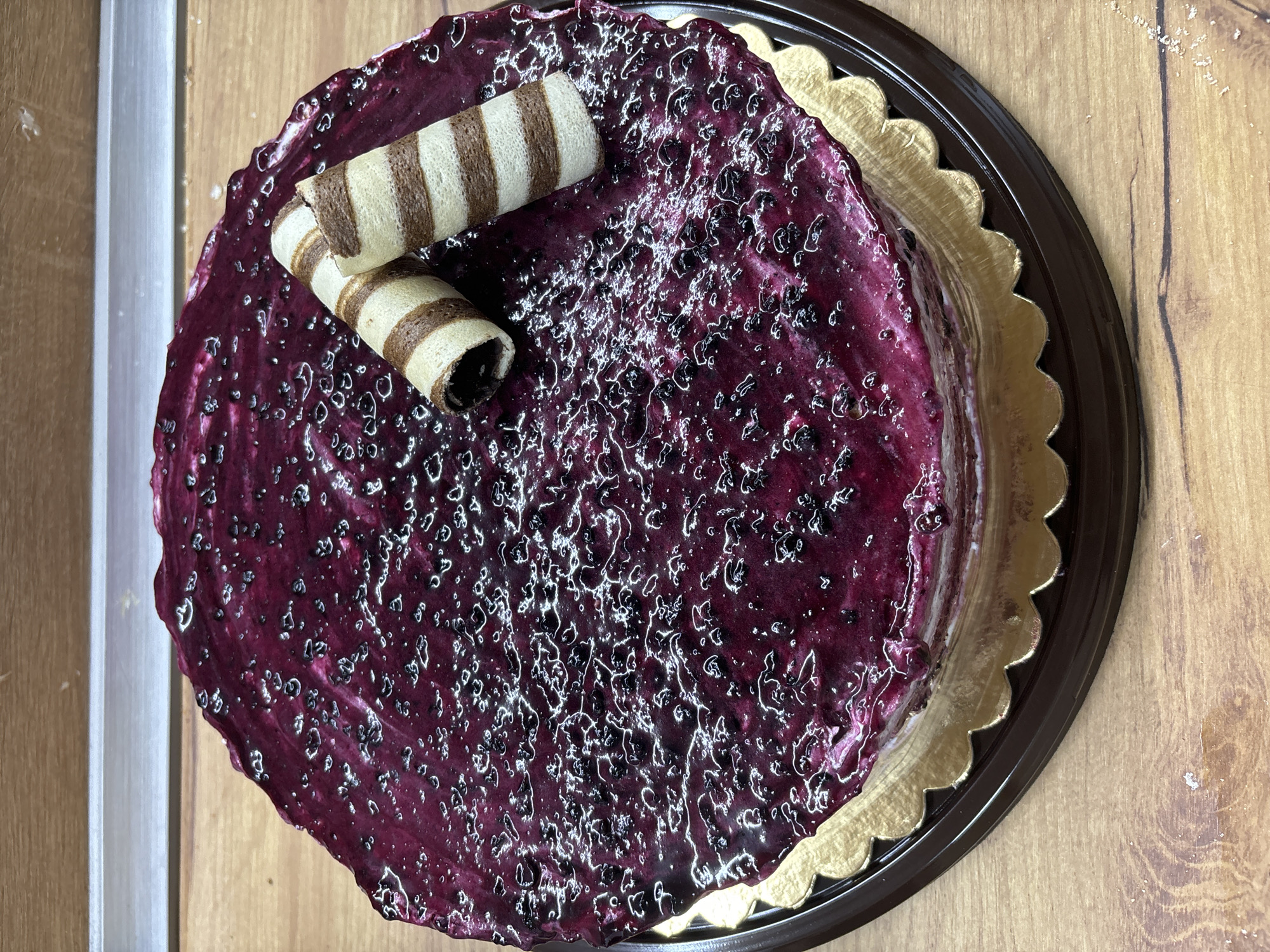 An overhead shot presents a round cake, lavishly coated in a vibrant, deep purple blueberry glaze that exhibits a glossy, somewhat textured surface with visible fruit pieces. Two striped wafer rolls, light in color with dark brown bands, are artistically placed atop the cake, serving as a decorative accent. The cake rests on a gold-colored, scalloped cake board, which in turn sits within a larger, dark brown plastic tray or base.
The entire arrangement is positioned on a light brown wooden surface, likely a table or countertop, revealing distinct wood grain patterns. To the left of the cake, a lighter vertical strip, possibly a wall or another piece of furniture, is visible. The lighting is bright, emphasizing the reflective quality of the glaze and the intricate details of the wafer rolls. There is no specific activity taking place, rather the cake is presented in a static display. No people are visible, and there is no discernible text in the image. The setting is indoors, with the specific location context being Malki Iskar, Bulgaria.