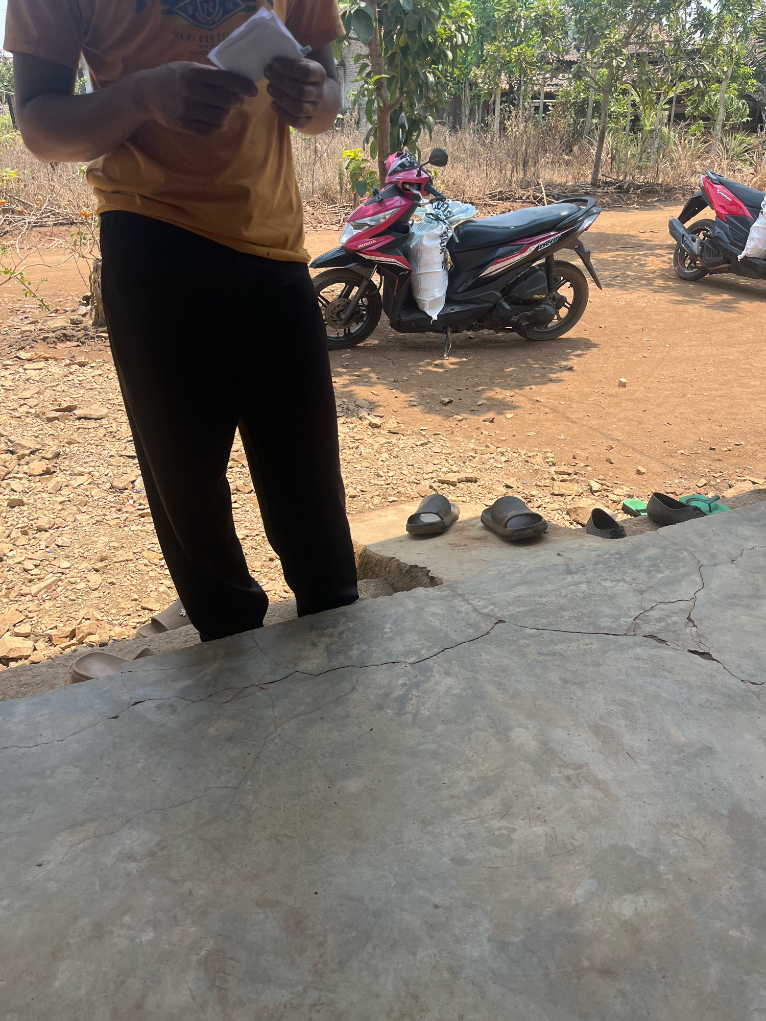 The image shows an outdoor scene with two motorcycles and a person standing in the foreground. The person is wearing a yellow t-shirt and black pants, and is holding a white object. There are several pairs of slippers and sandals on the ground near the person. In the background, there is a building with a thatched roof and trees. The ground is covered with dirt and small stones. The scene appears to be in a rural area of Penantian, Indonesia.
