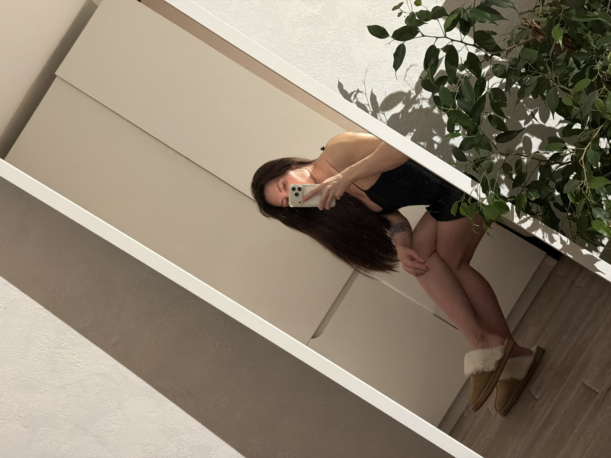 A woman is taking a selfie in a mirror. She is wearing a black top, and slippers with a fur trim. The reflection also shows a plant to the right of the woman, and a white wall with cabinetry. It is not possible to identify the specific location in Perm, Russia.
