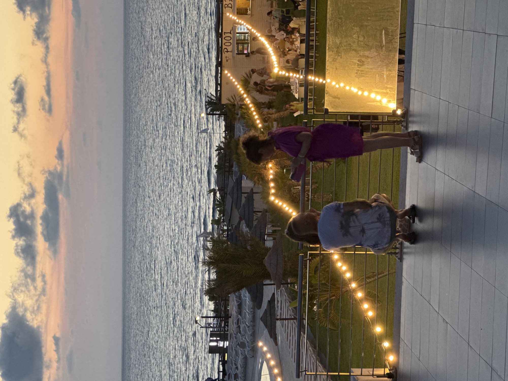 The media file captures a scenic evening view from a balcony overlooking the sea in Isola delle Femmine, Italy. The scene features two young girls in the foreground, one wearing a purple dress and the other a light blue top. The girl in the purple dress is looking down at her phone, while the girl in the blue top is crouched down, seemingly interacting with something on the ground. The balcony is adorned with a string of warm, glowing lights, creating an inviting atmosphere.
Beyond the balcony, the Mediterranean Sea stretches out, its surface rippling gently. The sky above shows a blend of soft twilight colors, with hints of orange and pink from the setting sun, interspersed with dark clouds. On the shore, beach umbrellas are visible, suggesting a daytime activity that has wound down for the evening. In the background, a building with "POOL BAR" printed on it can be seen, illuminated by more festive lights, hinting at a lively establishment. The overall setting evokes a peaceful and relaxed end to a day at the beach, with a touch of evening entertainment.