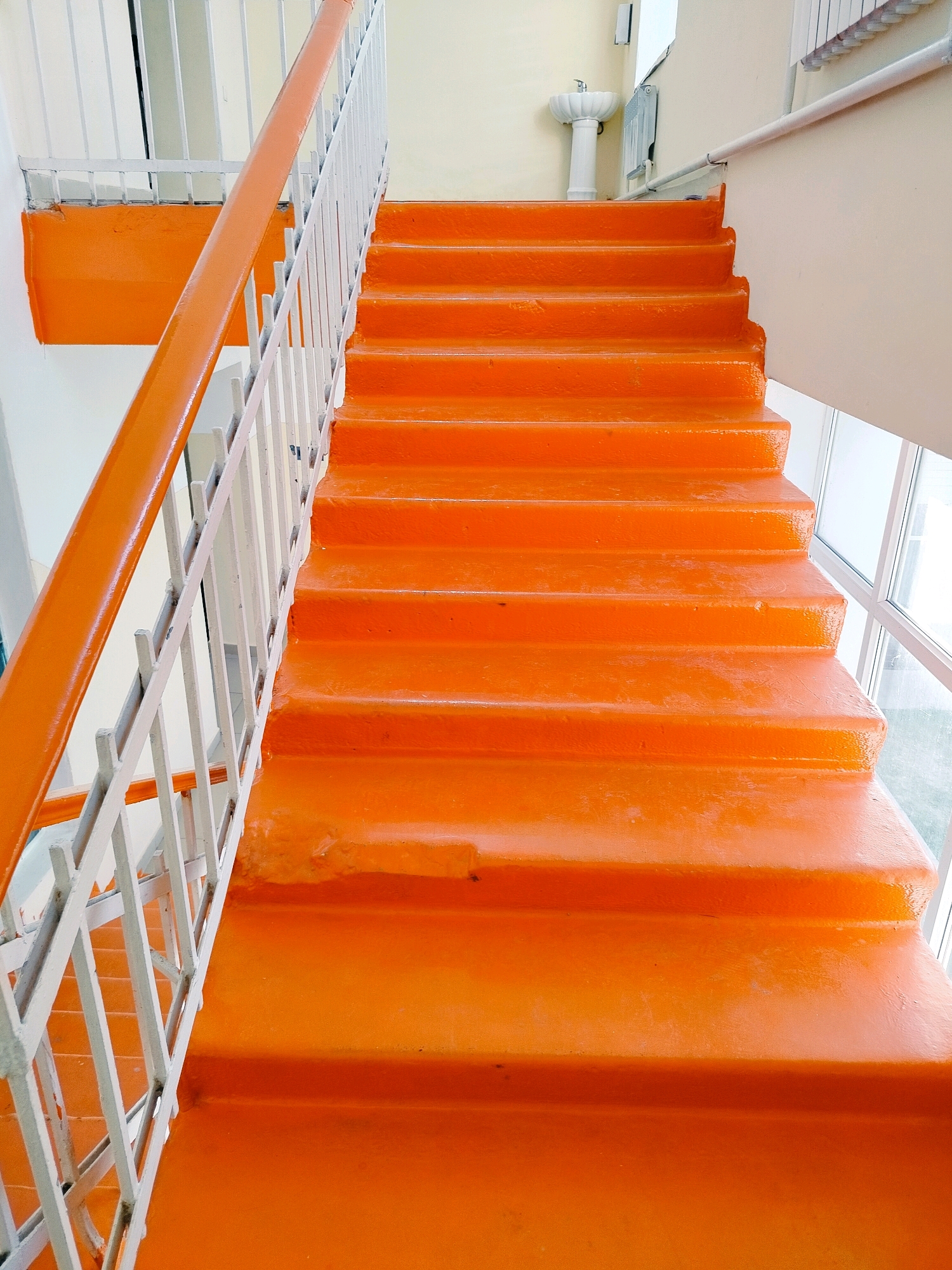 An indoor staircase with bright orange steps and an orange handrail. The staircase has a white railing on the left side and appears to be constructed of concrete. The walls are painted white and cream. In the background, there is a white pedestal sink and a radiator. The scene is well-lit, with natural light coming from a window on the right side of the image. The image appears to be taken in a public building or an apartment complex. No people are visible in the image. The location of Көлкент, Kazakhstan is provided as context for the image, but there are no specific visual cues in the image that directly indicate this location.