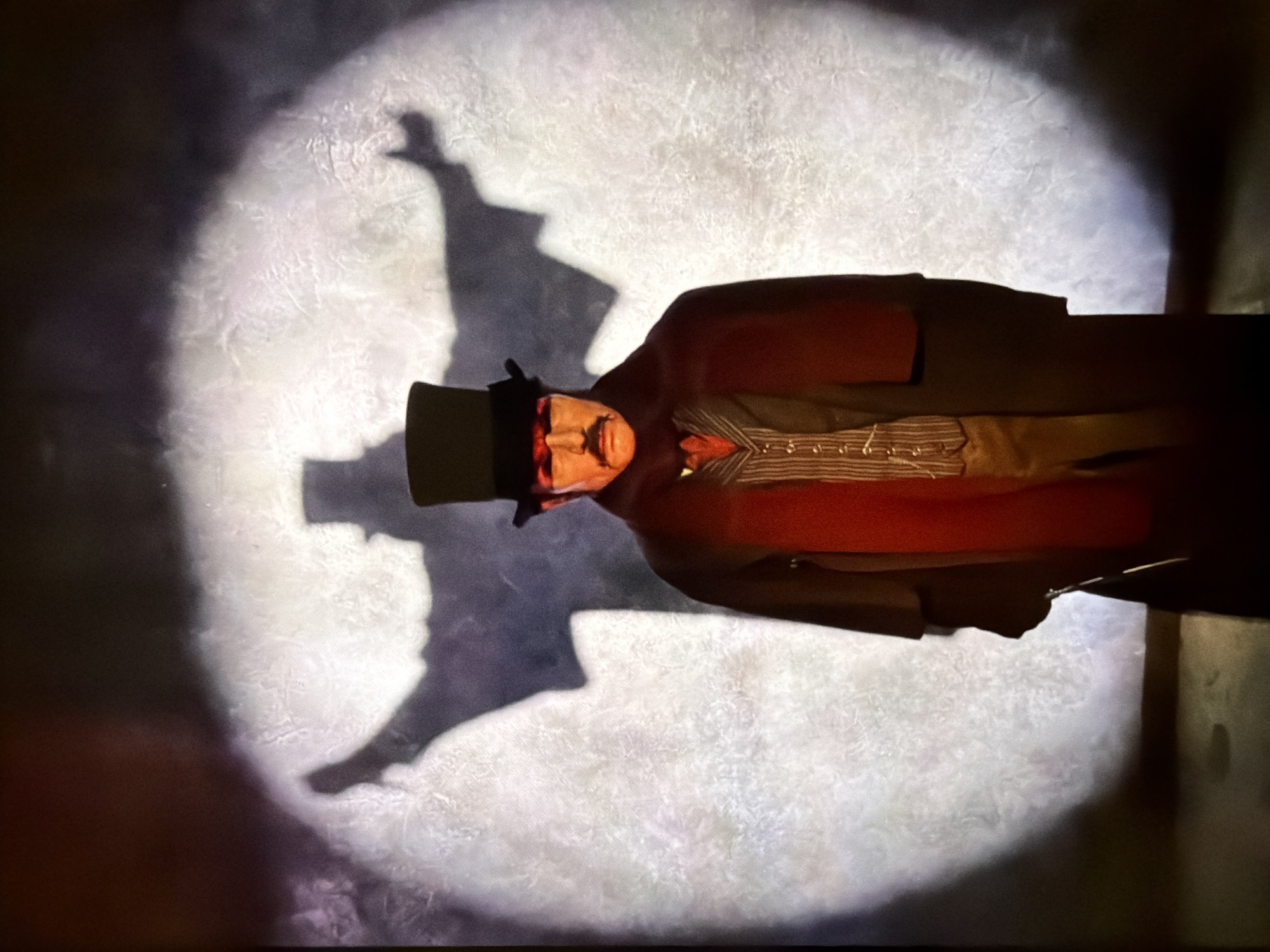 A man in period clothing (top hat, frock coat, striped vest) stands against a wall illuminated by a spotlight, which also casts a large shadow of him on the wall. The scene appears to be a theatrical presentation or display. The setting is likely London, United Kingdom, given the costume style.