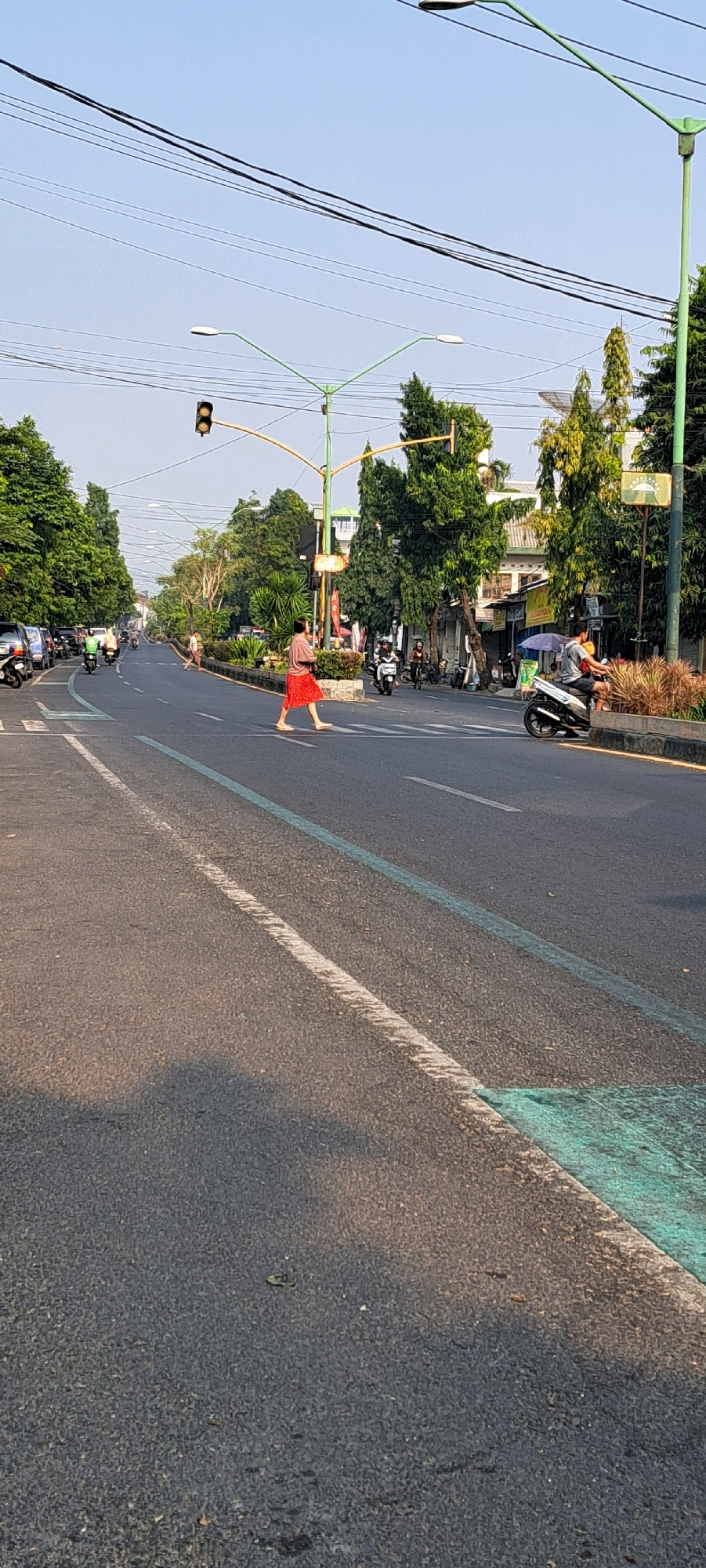 The image depicts a street scene in a city. A person in a red skirt is walking across a pedestrian crossing. There are several motorcycles and cars on the road, indicating traffic. Trees line the sides of the road, and buildings are visible in the background. Streetlights and traffic signals are also present, suggesting an urban environment. The sky is clear and blue, and the overall lighting suggests it is daytime, likely morning or afternoon. The scene appears to be a typical street in a built-up area.