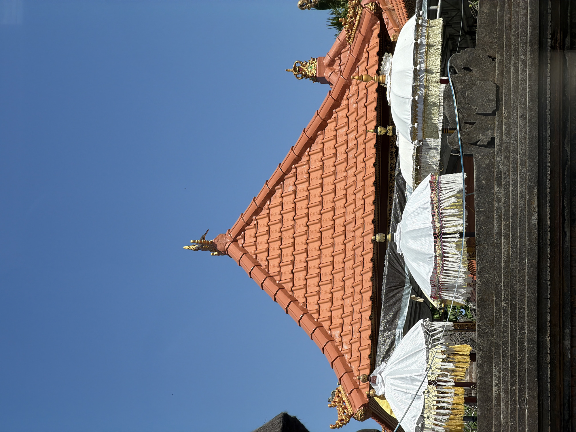 The image depicts the exterior of a traditional Balinese building with an orange-tiled roof, adorned with gold-colored decorative elements, under a clear blue sky. In the foreground, several white ceremonial umbrellas with ornate golden trim and fringes are displayed. These umbrellas are positioned on top of a stone structure. The overall scene suggests a cultural or religious setting, likely a temple or a place of ceremony. The image does not contain any people. The scene is set outdoors. The exact location in Kediri, Indonesia, cannot be determined from the image alone, as it lacks any specific landmarks or identifying features of that city.