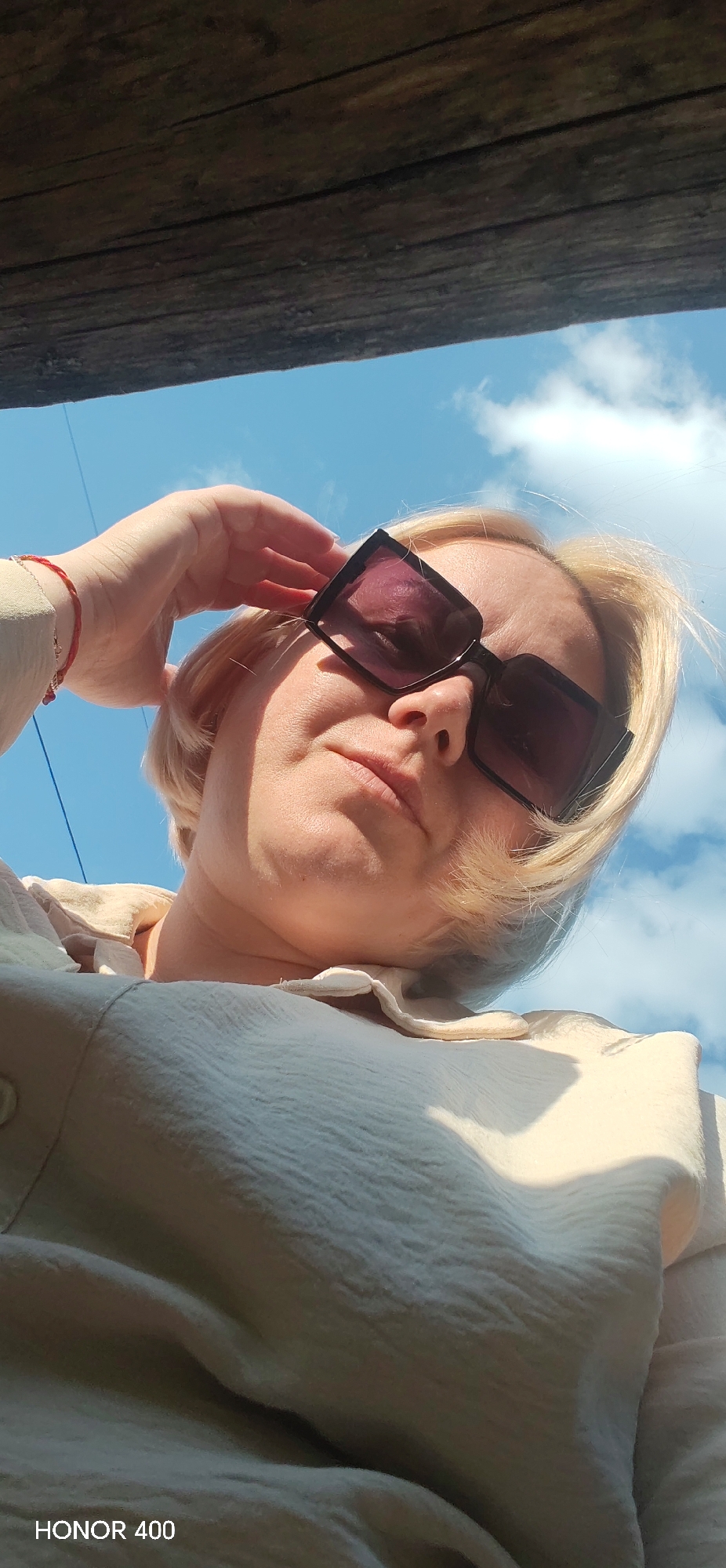 A low-angle shot shows a woman with short, blonde hair and large, dark sunglasses. She is touching the side of her glasses with her left hand, which has a red bracelet on the wrist. She is wearing a light-colored shirt. Above her, a blue sky with some clouds is visible, along with a dark, wooden structure. The photo was apparently taken with an HONOR 400. The location of Noviy, Russia cannot be determined from the image.