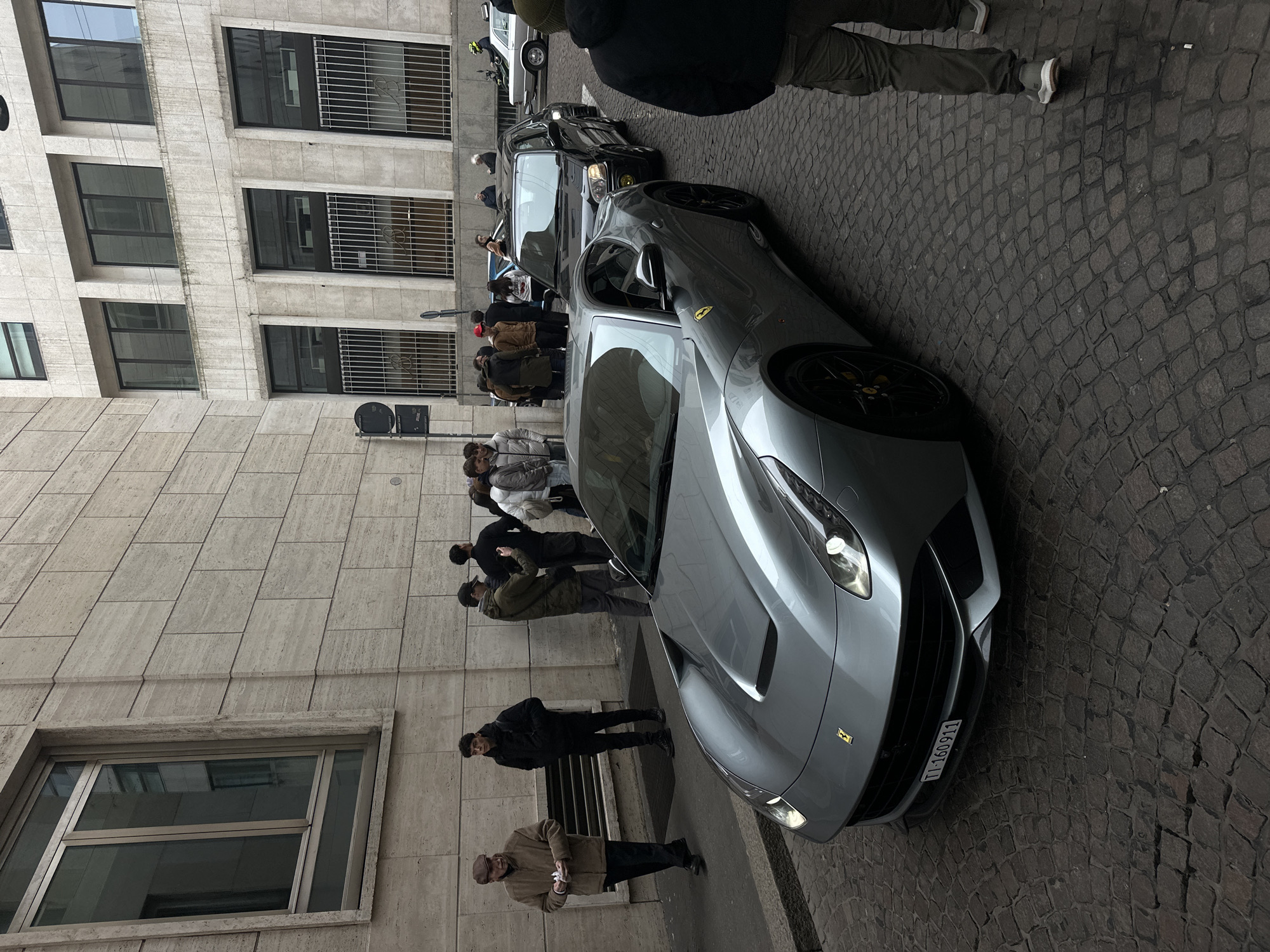 The image depicts an overcast street scene in Milan, Italy, characterized by a grey Ferrari F12berlinetta positioned centrally on a dark grey cobblestone street. The car faces left, with its headlights illuminated and a Swiss license plate, "TI-160911," displayed on its front bumper. Behind the Ferrari, a black hatchback or small SUV is visible, followed by a silver sedan further down the street on the right, and a motorcycle/scooter parked on the far right.
On the left, a stone-clad building with prominent, vertically oriented rectangular windows and a smaller street-level window defines the backdrop. A sidewalk runs along this building. A group of approximately ten individuals is clustered on the sidewalk and near the vehicles. Actions include observing the cars, conversing, and one person (wearing a green jacket and cap) appears to be photographing the scene with a smartphone. Two men are walking on the sidewalk on the left: one older man in a brown jacket and flat cap holds a small white object, moving left; another younger man in a black jacket walks left. In the foreground, the lower half of a person wearing dark cargo pants and light grey sneakers is visible on the right, walking right, partially obscuring the view of the street. The buildings exhibit a beige-grey stone facade, with some upper windows featuring dark frames and others at street level protected by white metal grates. The overall environment suggests an urban setting with historical architectural elements.