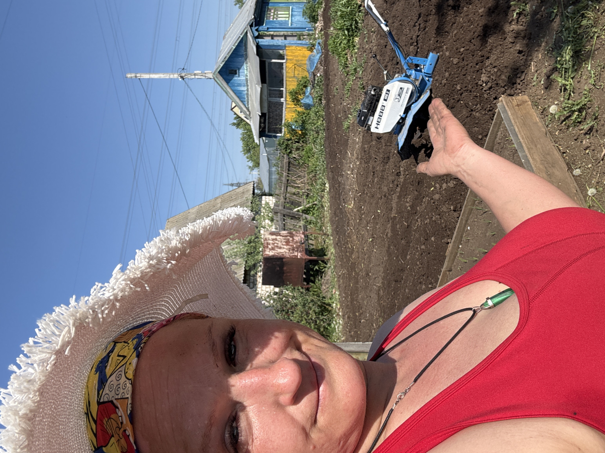 A selfie-style image shows a woman wearing a white, fringed hat and a red tank top. She has a colorful headscarf underneath the hat and is wearing a necklace with a green pendant. Her hand is partially visible, extended towards a small blue and white tiller or cultivator machine working in the soil. Behind her, there is a rural scene with a weathered wooden house painted in blue and yellow. In the background, power lines stretch across a bright blue sky. A shed is also visible. The location is likely a garden or plot of land near a residential area in Chaikovsky, Russia, based on the architectural style of the house and the agricultural activity.