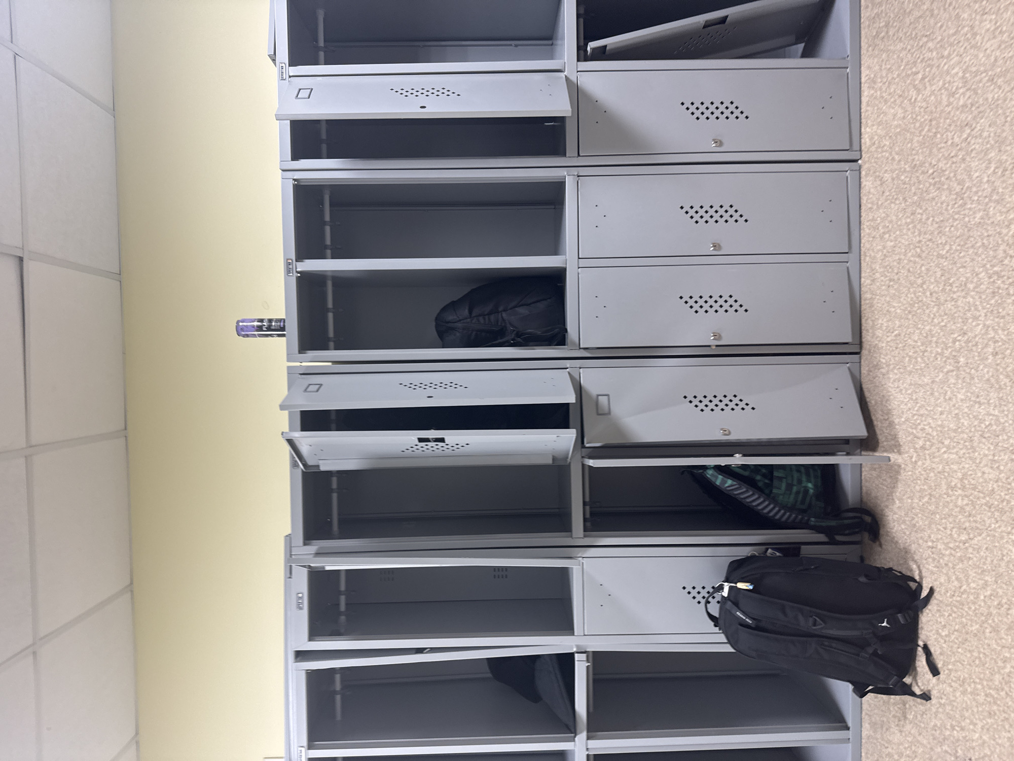 The image depicts a set of gray metal lockers in Kazan, Russia, positioned against a light yellow wall and a beige carpeted floor. The lockers are stacked vertically, with some doors open and others closed. Several lockers contain items, including black and green backpacks. One locker contains a purple bottle. The lighting is moderate, casting shadows within the lockers.