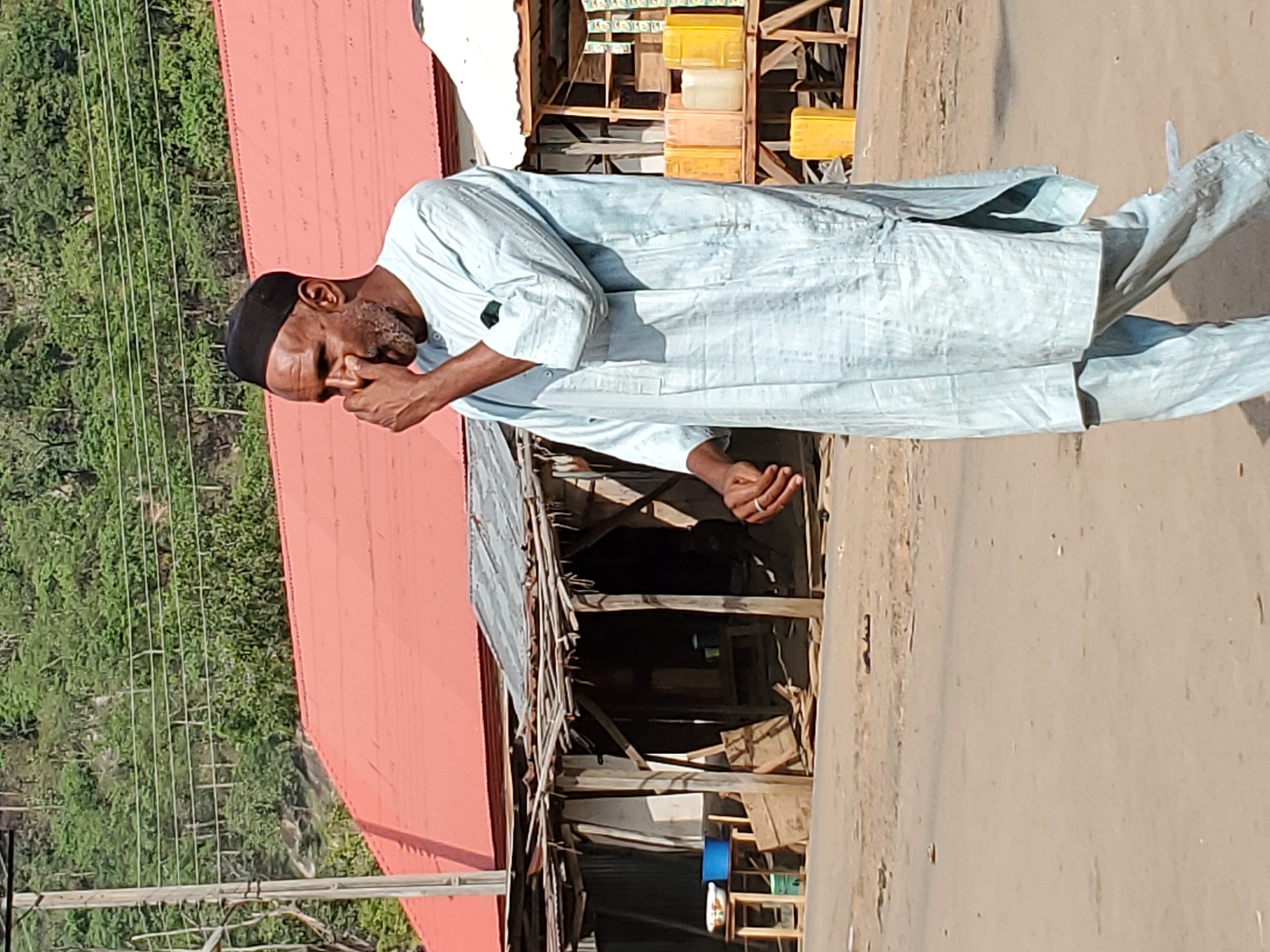 A full-body image captures a man walking or standing outdoors on what appears to be an unpaved street or path in Mararaba, Nigeria. The man, seemingly middle-aged, is dressed in a light blue, long-sleeved tunic (possibly a thobe or kaftan) and matching trousers, topped with a black cap (kufi). He has a beard and is depicted with his right hand raised to his face, covering his mouth and part of his nose, suggesting a gesture of thought, discomfort, or wiping his face. He wears a ring on the ring finger of his left hand, which is slightly open.
Behind the man, on the right side of the frame, there is a rustic, makeshift market stall or shop, constructed from wooden beams and corrugated metal sheets, displaying various yellow and white plastic containers. To the left, a building with a prominent red corrugated iron roof is visible. The background features lush green trees and foliage, indicating a natural or semi-rural environment, with multiple utility wires strung across poles. The ground is dusty and uneven.
The bright sunlight suggests it is daytime, under clear weather conditions. The scene portrays an ordinary, everyday moment in a local community setting. There is no visible text in the image.