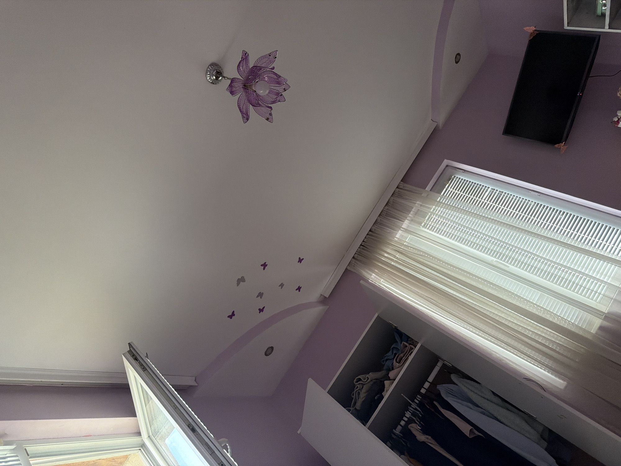 This upward-angled interior shot, taken in Garmen, Bulgaria, showcases a room, likely a bedroom, with a distinctive lavender and white color scheme. The white ceiling features a decorative architectural arch, painted in a soft lavender or light purple, adorned with several small, purple butterfly decals. A prominent purple, flower-shaped chandelier with a single bulb hangs from the white ceiling, complementing the room's color palette. A recessed light fixture is visible within the lavender ceiling arch.
Bright daylight streams into the room through a large window on the right, which is fitted with horizontal blinds and sheer white curtains. Below this window, an open white wardrobe or closet reveals various clothes, some folded and some hanging on racks. Another window, partially open, is visible on the lower left. A black flat-screen television, decorated with small corner embellishments, is mounted on the light purple wall. The overall ambiance is bright and serene, suggesting it is daytime. No people or visible text are present, and no specific activity is occurring.