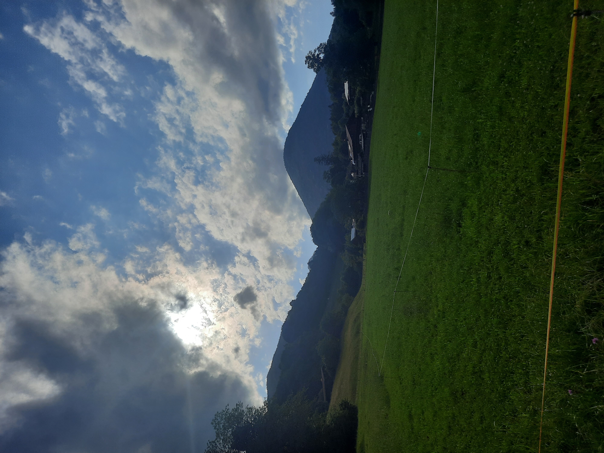 A wide-angle, vertical shot captures a lush, green meadow stretching towards rolling hills under a dramatic sky. The sun, partially obscured by dark, textured clouds, peeks through, casting a soft light on the scene. The sky is a mix of deep blue and wispy white clouds, with patches of bright sunlight.
In the mid-ground, a cluster of houses with dark roofs are nestled amongst trees and on the slopes of the hills. The hills themselves are covered in dense, dark green foliage, creating a silhouette against the brighter sky. The foreground is dominated by the vibrant green grass of the meadow, with a thin, white electric fence running horizontally across it. A hint of a yellow cord is visible at the very bottom of the frame. The overall impression is one of a tranquil, rural landscape bathed in dynamic, natural light.