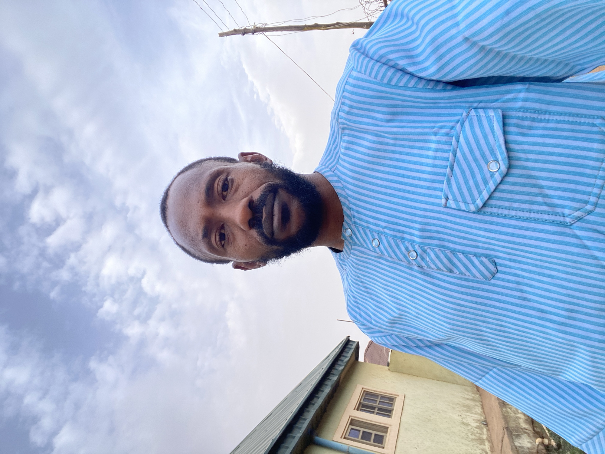 A low-angle shot captures a man with a beard and mustache, wearing a blue and white striped collared shirt with a buttoned pocket. His face is directed towards the camera. The background features a cloudy sky, utility poles with wires, and the top of a building with two windows. Based on the prompt, the location is in Sabon Tasha, Nigeria.