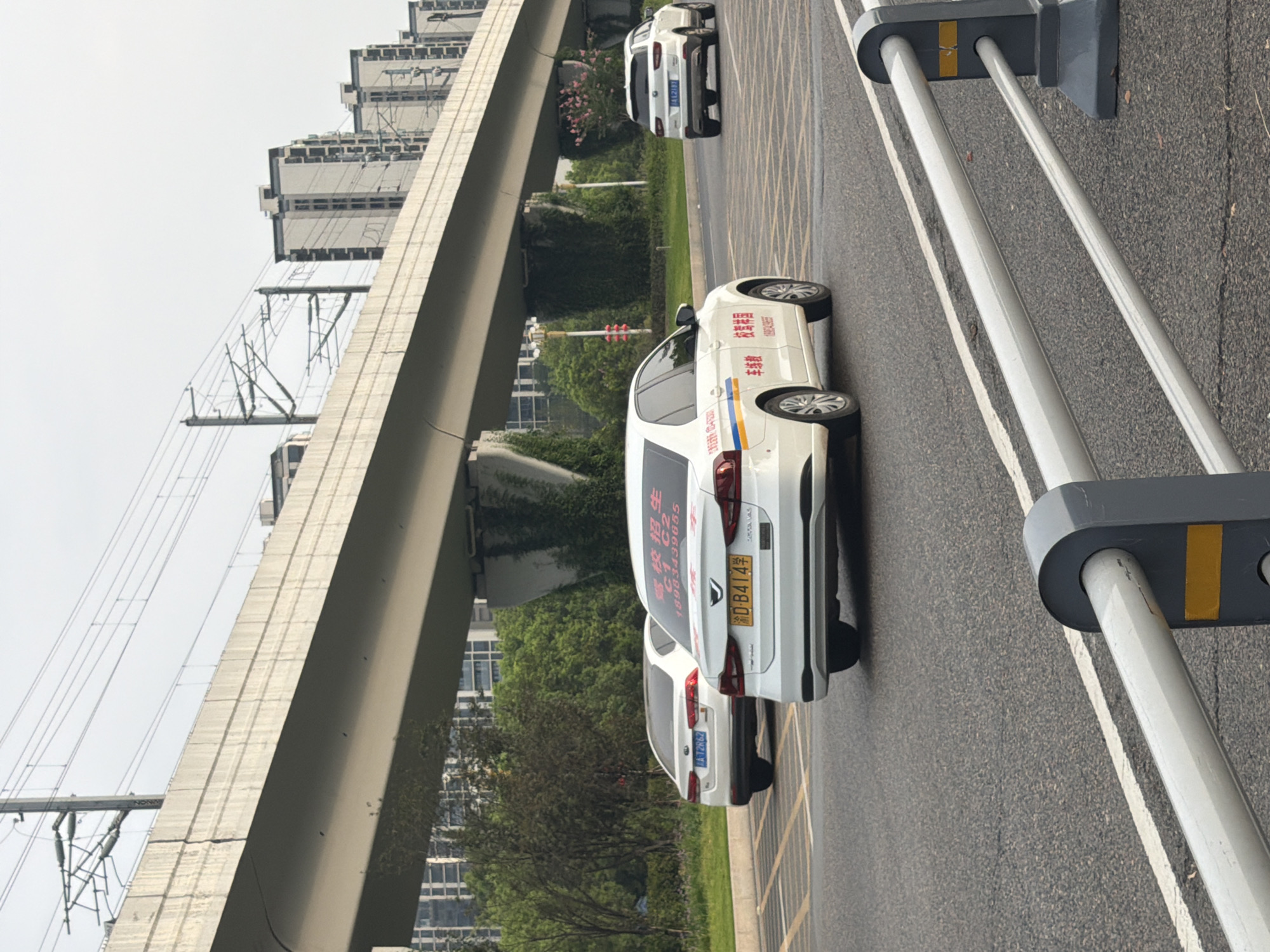 The image captures a street scene in Jiangjin, China, beneath an elevated roadway. In the foreground, a gray and silver metal barrier with reflective yellow markings is partially visible. The road surface is asphalt, with visible lane markings. Three white cars are present. The closest car, a sedan, is facing away from the viewer and has Chinese characters and a license plate "D-B414" on its rear. A second white sedan, also facing away, is to the left and has a license plate "A-12R62". Further down the road, a white SUV is visible from the rear, with a license plate that appears to read "AA2181". In the background, trees and tall residential buildings line the sides of the road, and the elevated roadway, supported by concrete pillars, extends into the distance. Electrical wires for an overhead train system are visible above the roadway. The sky is overcast.