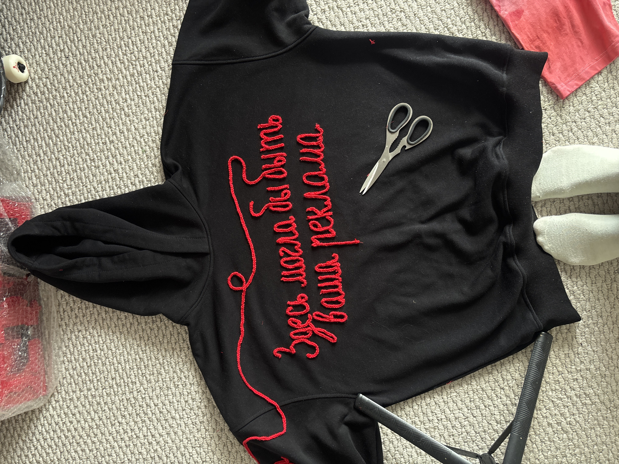 A black hooded sweatshirt with red embroidery in Cyrillic script reading "Здесь могла бы быть ваша реклама" ("Your advertisement could be here") is laid out on a light-colored carpet. A pair of silver scissors rests on the hoodie. Nearby are a red shirt, white socks, and the wheel of a skateboard. Parts of a chair and packaging are also visible. The location is unknown.