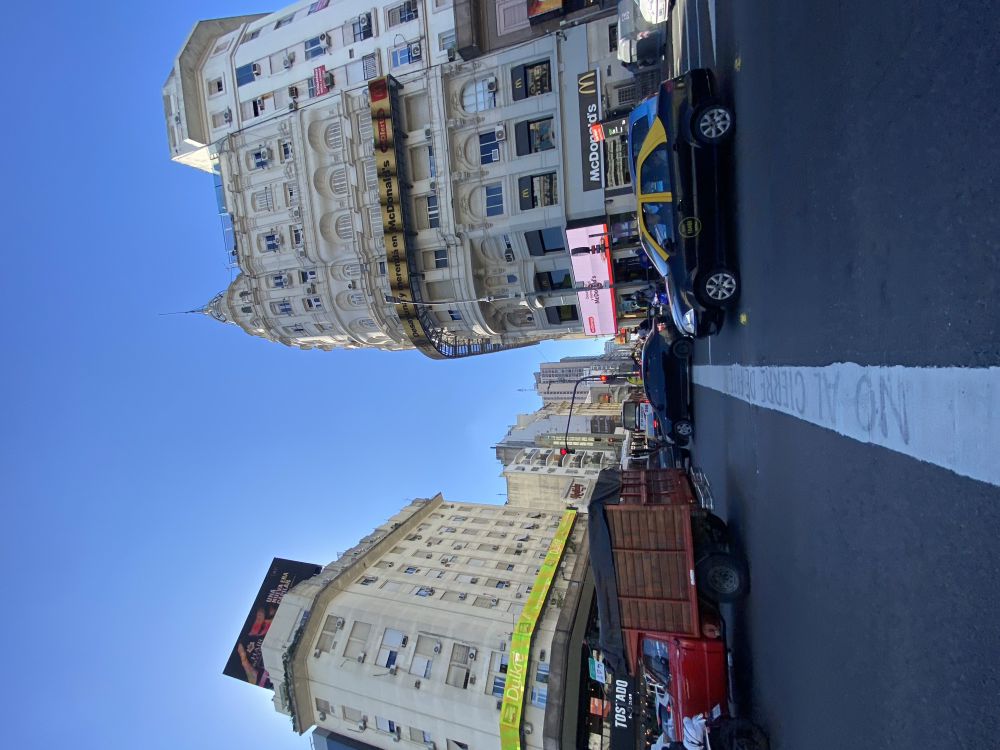 The image captures a street scene in Buenos Aires, Argentina. Two prominent buildings face each other across a city street. One building features a rounded facade with ornate architectural details and a McDonald's restaurant on the ground level, with signage visible. The other building displays large advertising billboards for "UNA NUEVA ERA CAPILAR." A taxi and a red delivery truck are visible in the street. The street is marked with a solid white line, and the words "NO AL CIERRE DE" are painted in white on the road. The sky is blue.
