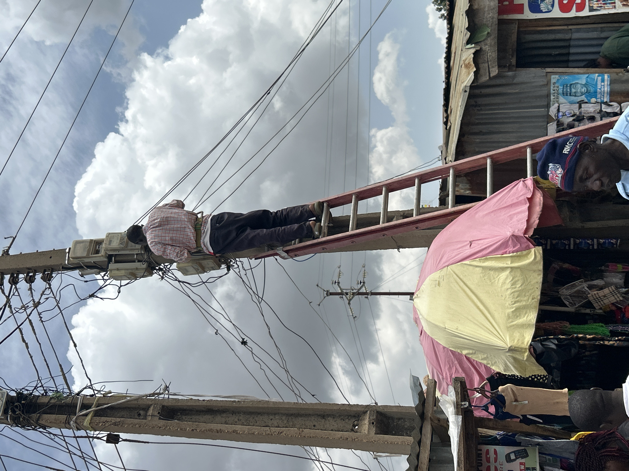The image captures a bustling outdoor scene, likely a market or commercial street, in a developing urban area during the daytime under a partly cloudy sky. The central focus is a man in a plaid shirt and dark pants, actively climbing a red ladder that leans against a utility pole. He appears to be engaged in work related to the intricate and dense network of electrical wires and meters attached to the pole. The wiring appears haphazard and numerous, suggesting informal electrical connections.
To the right, partially visible, is another man wearing a blue beanie that reads "RMCC" and a light blue top, looking downwards. A large pink and yellow umbrella is deployed, possibly providing shade for a nearby market stall. Behind the umbrella and the second man, parts of market stalls are visible, constructed with corrugated metal and wood, displaying various goods such as textiles, sunglasses, and general merchandise. A blue and white poster on one of these stalls shows a portrait of a man and the partial text "SELFLESS SERVICE." Near the bottom right, a small sign featuring an older model mobile phone and the word "RECHARGE" is discernible, indicating a service point. The overall scene conveys a sense of industrious activity and the challenges of infrastructure in a crowded setting.